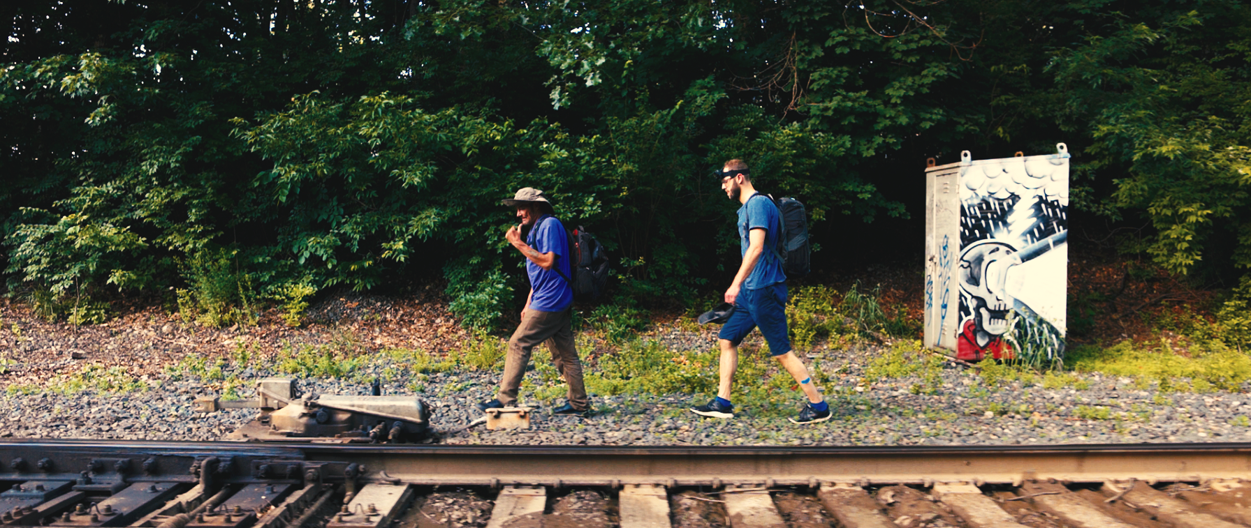 Two men walking along a railway track next to a graffiti-covered electrical box, with dense green trees in the background.