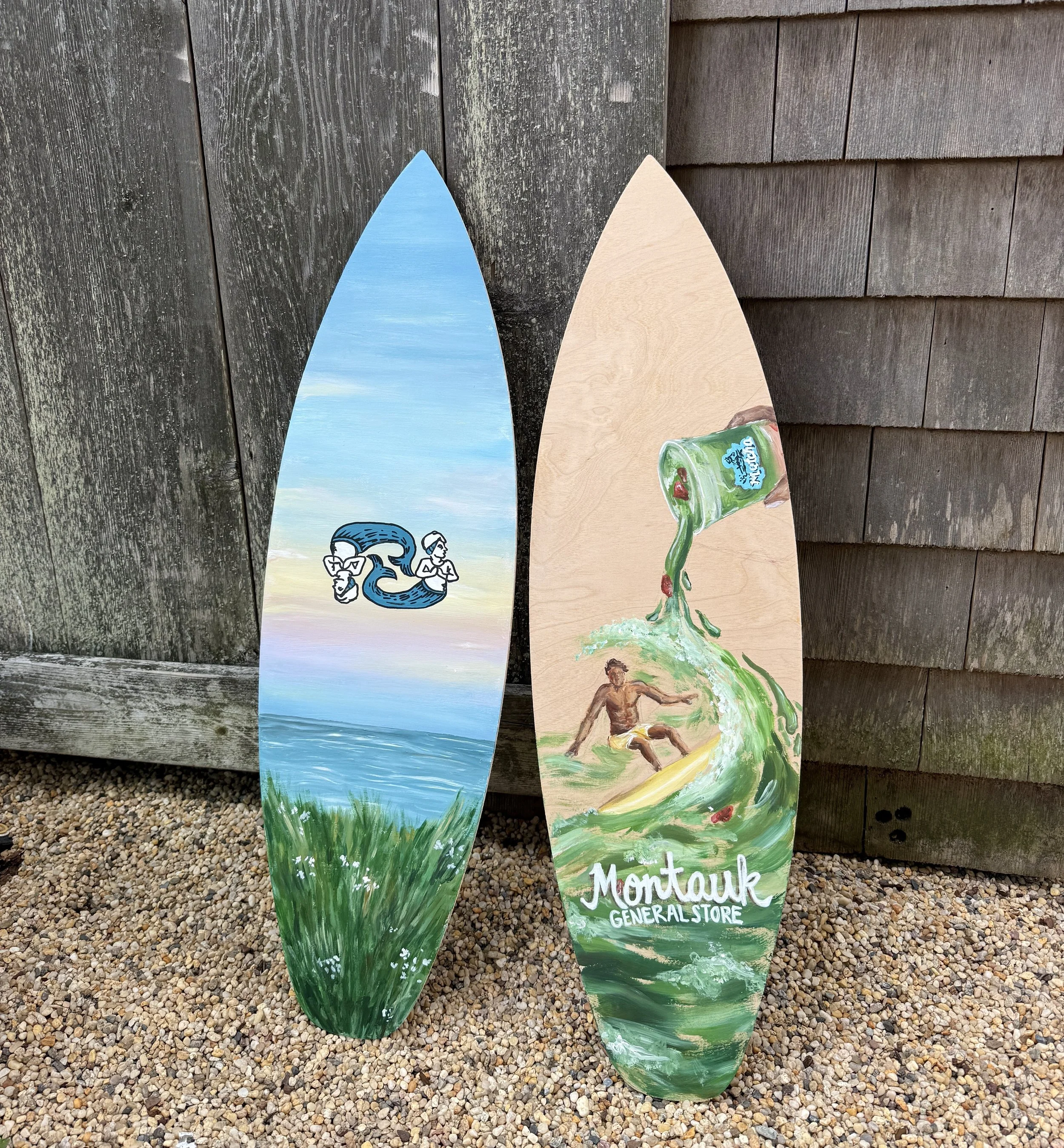 Painted wooden boards with ocean and landscape scenes, propped against a weathered wooden wall with shingles, on a gravel surface, with a logo for Montauk General Store at the bottom.