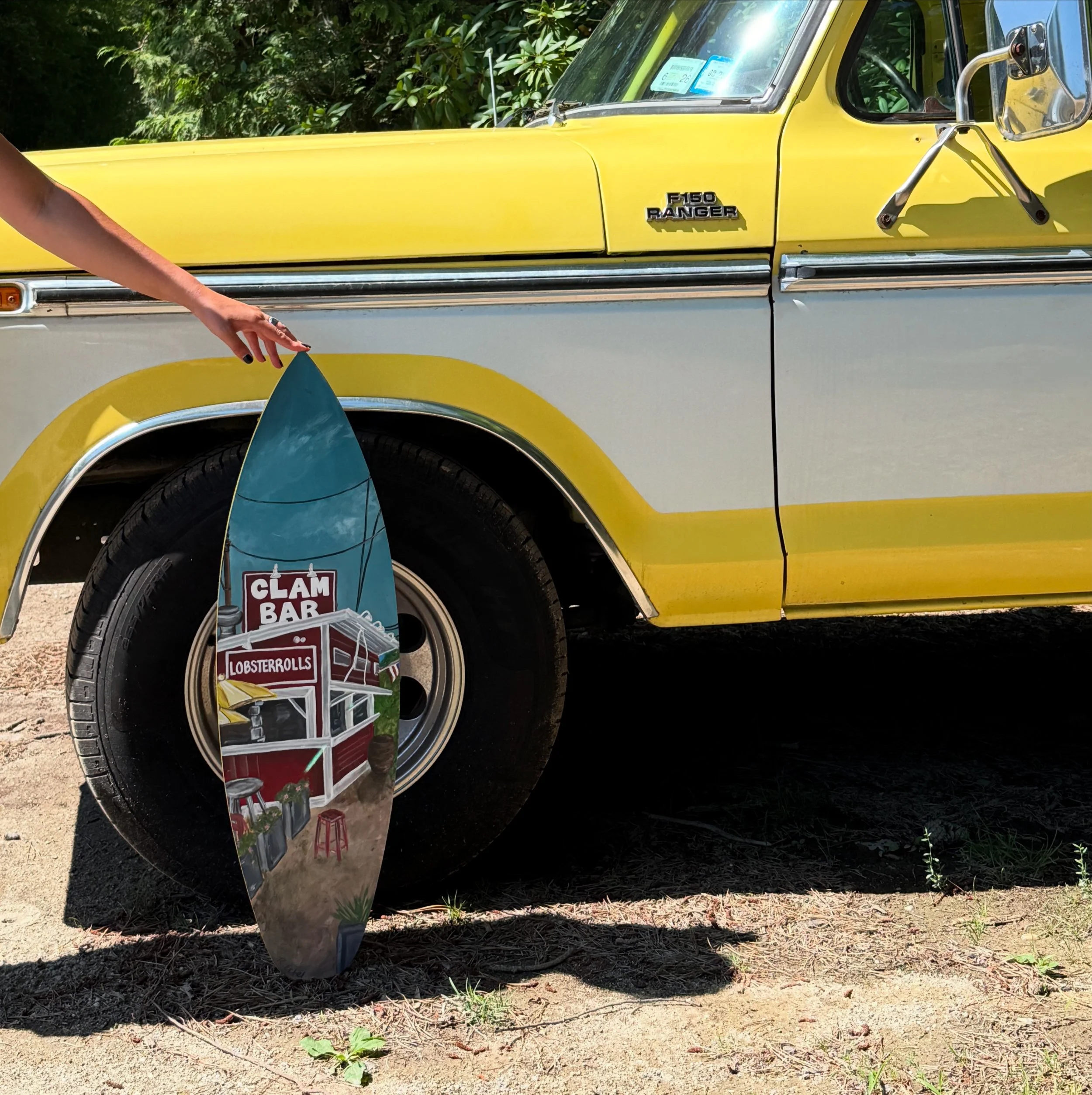A yellow vintage Ford F-150 Ranger pickup truck parked outdoors on a dirt surface with greenery in the background. Part of a person's arm is visible, and they are reaching towards a colorful surfboard or sign leaning against the truck, which advertises clam bars, lobster rolls, and other seafood.