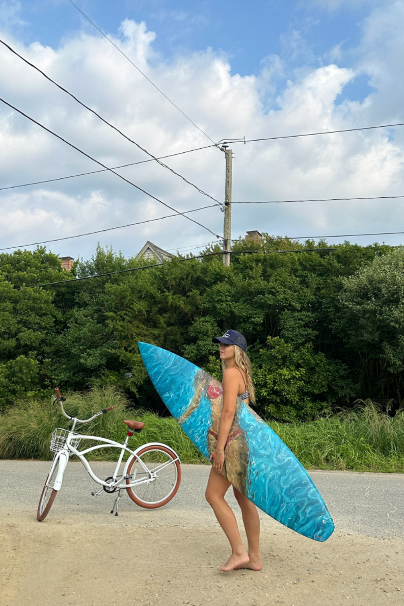 A woman in a swimsuit, baseball cap, and sunglasses holding a surfboard, standing barefoot next to a white bicycle with a brown seat on a dirt road with green bushes and trees in the background under a partly cloudy sky.