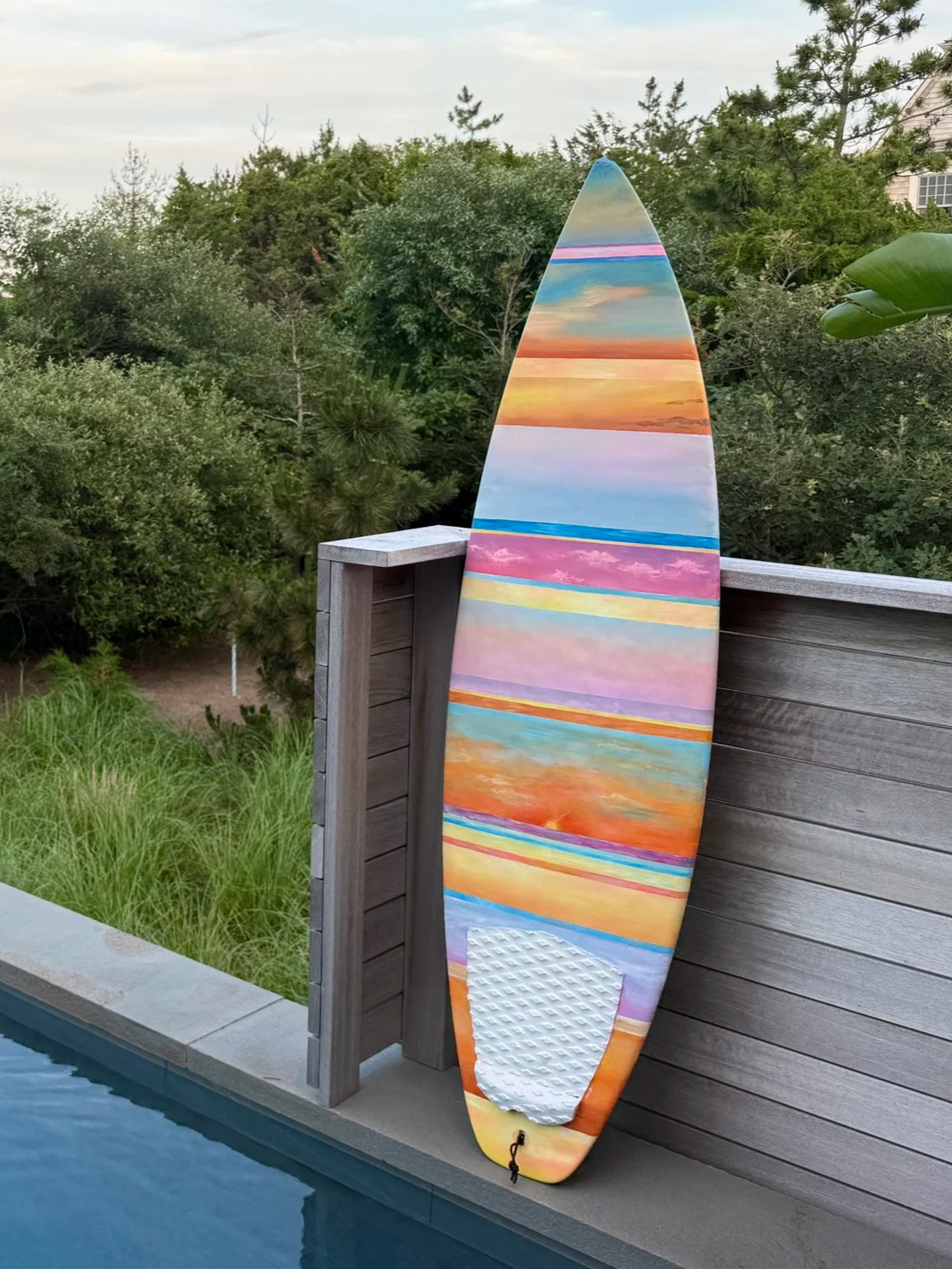 Colorful surfboard with a sunset landscape painted on it, leaning against a wooden fence beside a swimming pool.