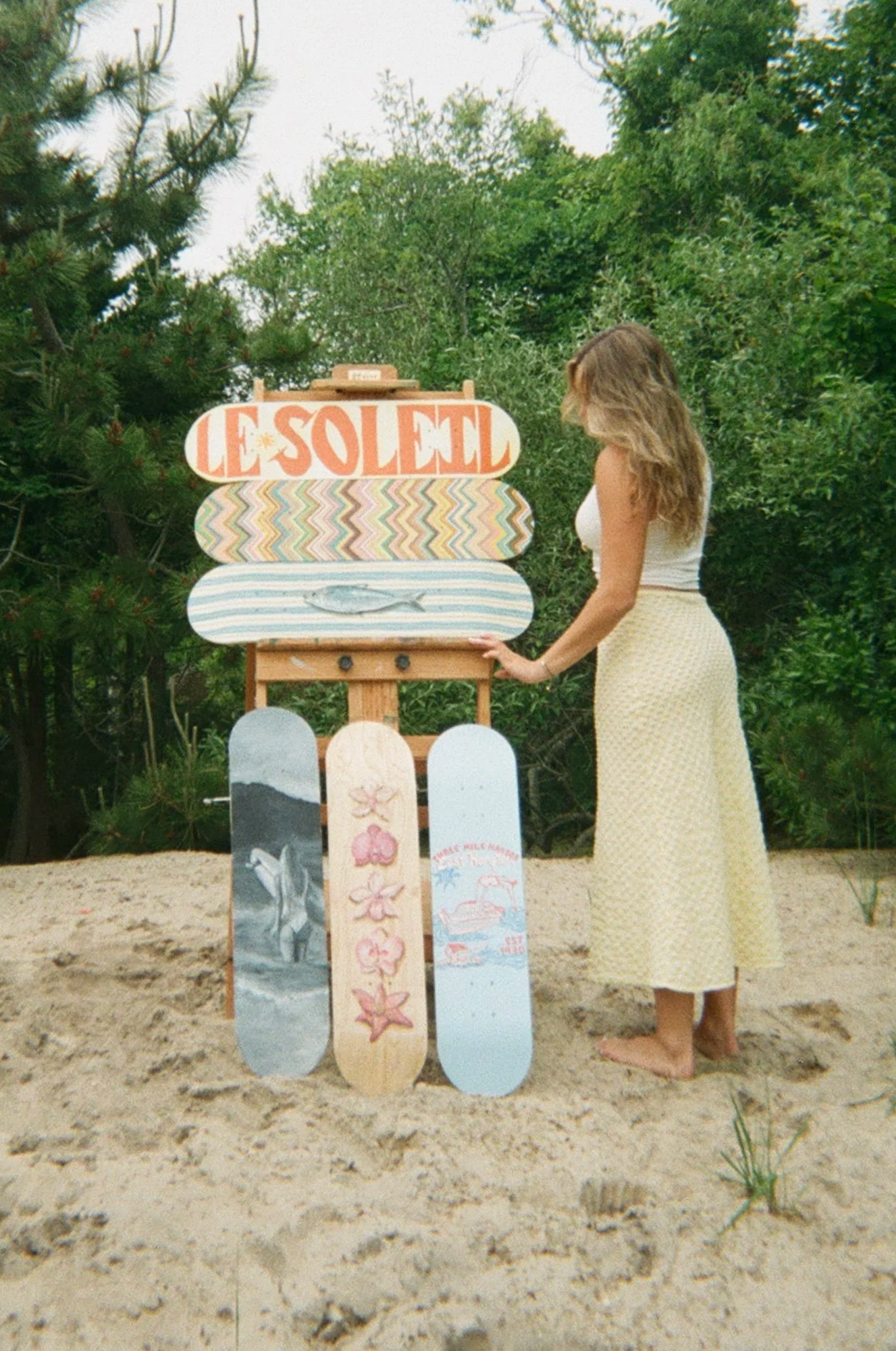 A woman in a yellow dress with white top holding onto a skateboard with a fish painting, standing on sandy ground surrounded by greenery and an island-themed sign that reads 'Le Soled' with three more skateboards with colorful designs leaning against