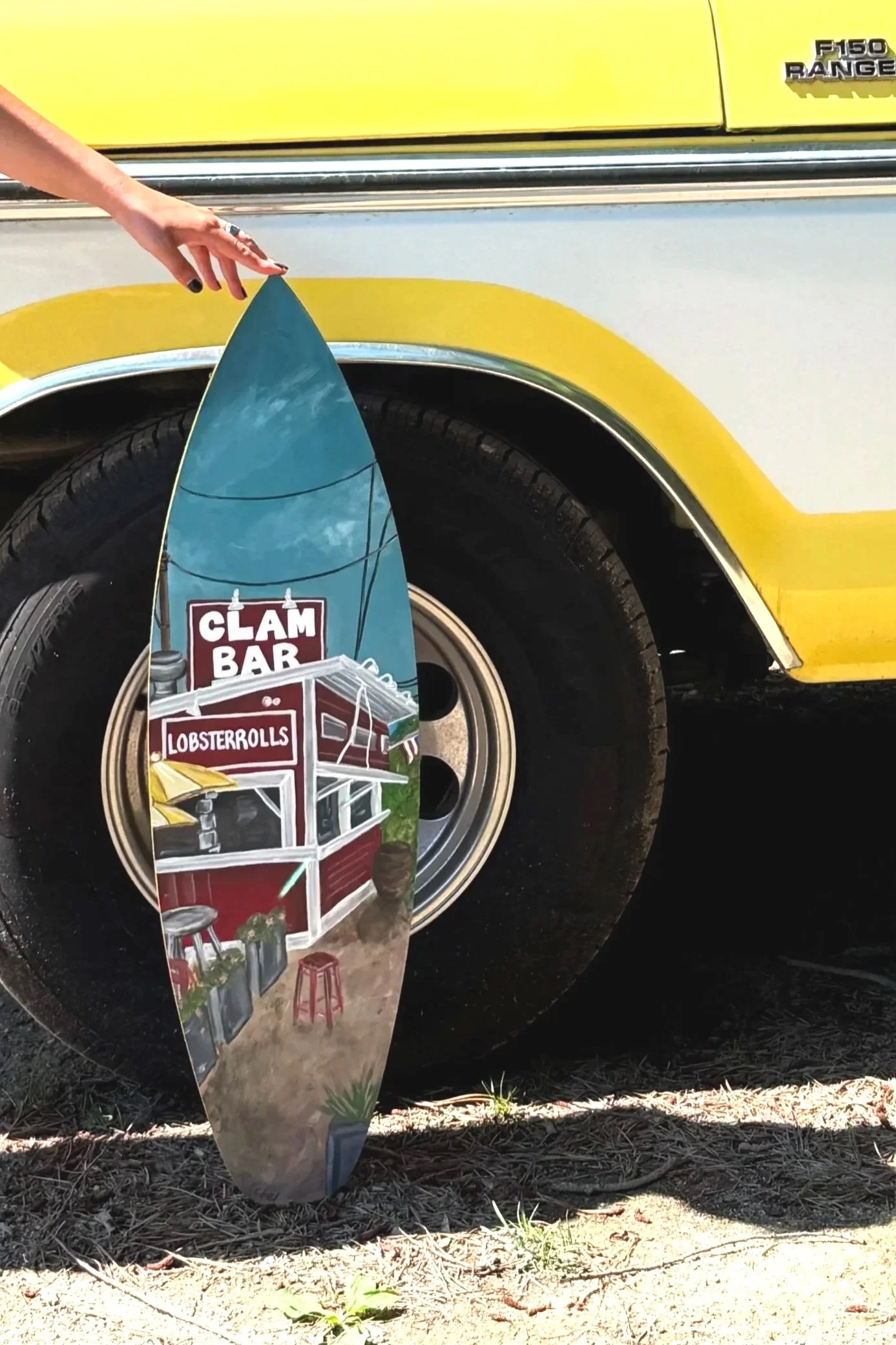 A yellow Ford F-150 Ranger truck parked on grass and gravel with a hand reaching towards a surfboard that has a painted illustration of a seafood stand with a sign reading "Clam Bar," "Lobster Rolls," and other seafood items.