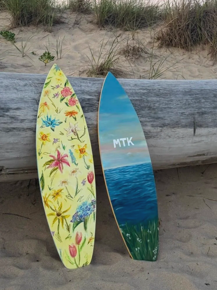 Two surfboards leaning against a wooden beam on a sandy beach with grass and sand dunes in the background. One surfboard is decorated with colorful floral designs, and the other features a painted ocean scene with the initials 'MTK'.