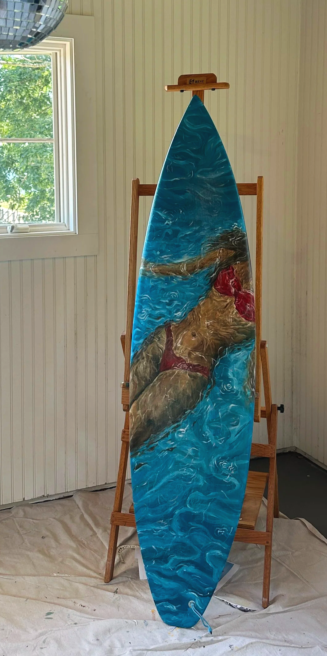 A painting on a surfboard featuring a woman in a red bikini with brown hair, swimming in a blue ocean background. The surfboard is displayed on a wooden easel indoors near a window with green foliage outside.