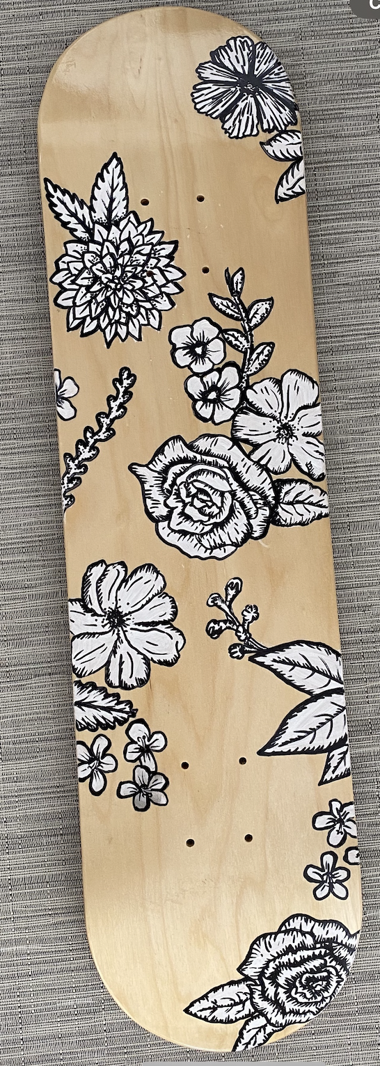 A skateboard deck with a natural wood finish decorated with black and white floral illustrations, including roses, hibiscus, and other flowers.