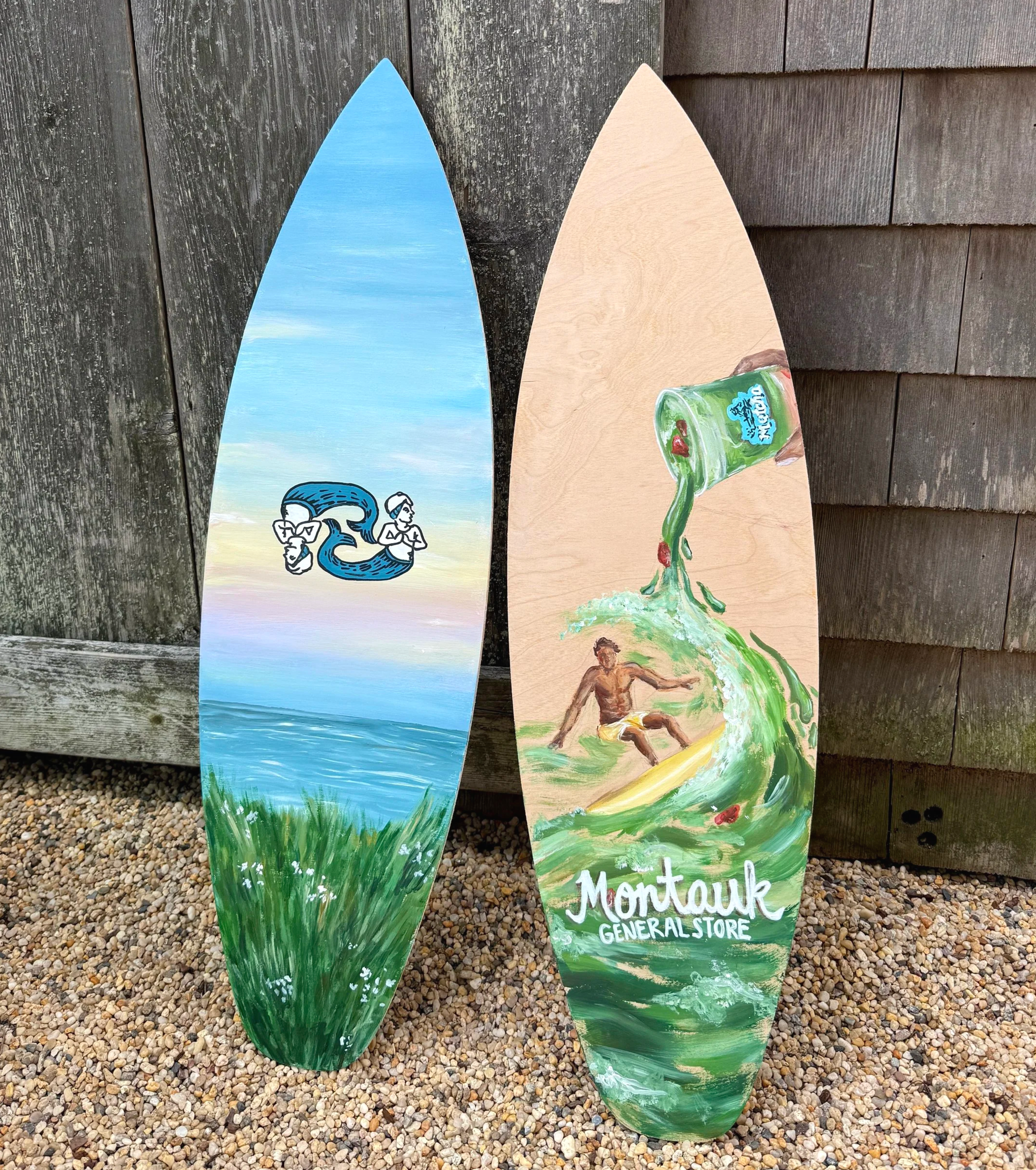 Painted wooden signs shaped like surfboards with scenic and advertising artwork, leaning against a weathered wooden fence on a gravel surface. One features a landscape with a blue sky, waterfall, and a logo; the other depicts a man sitting on the grass with a green bottle, with text that reads 'Montauk General Store'.