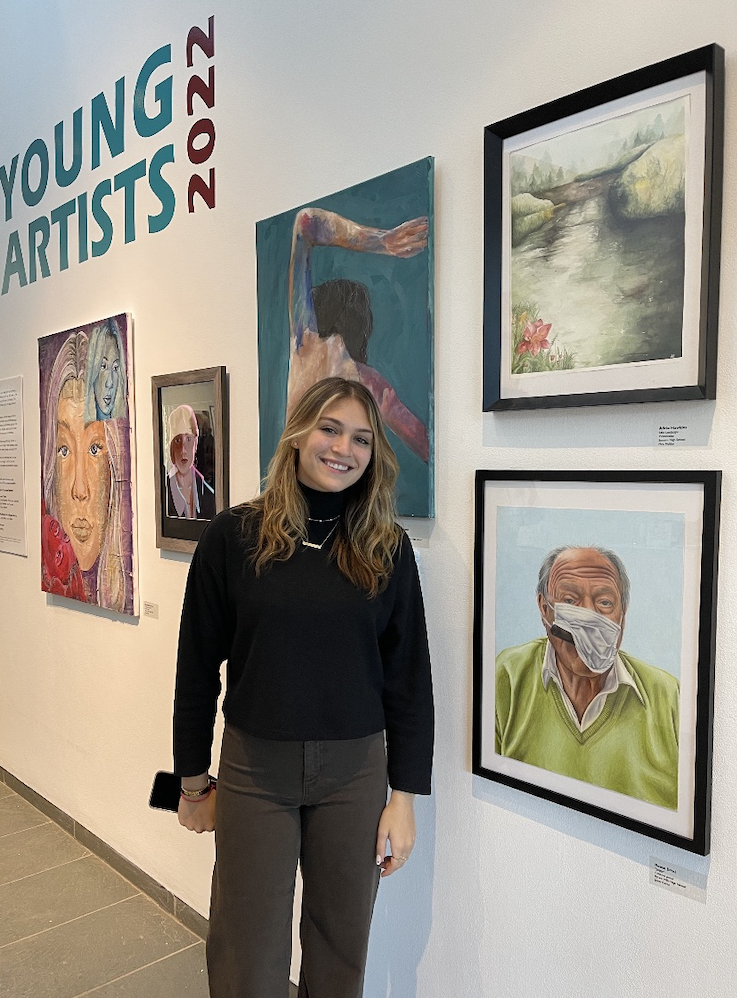 A young woman with long, wavy light brown hair, wearing a black sweater, gray pants, and holding a phone, smiling at an art exhibit with various paintings on the wall, including a portrait of an elderly man wearing a face mask.
