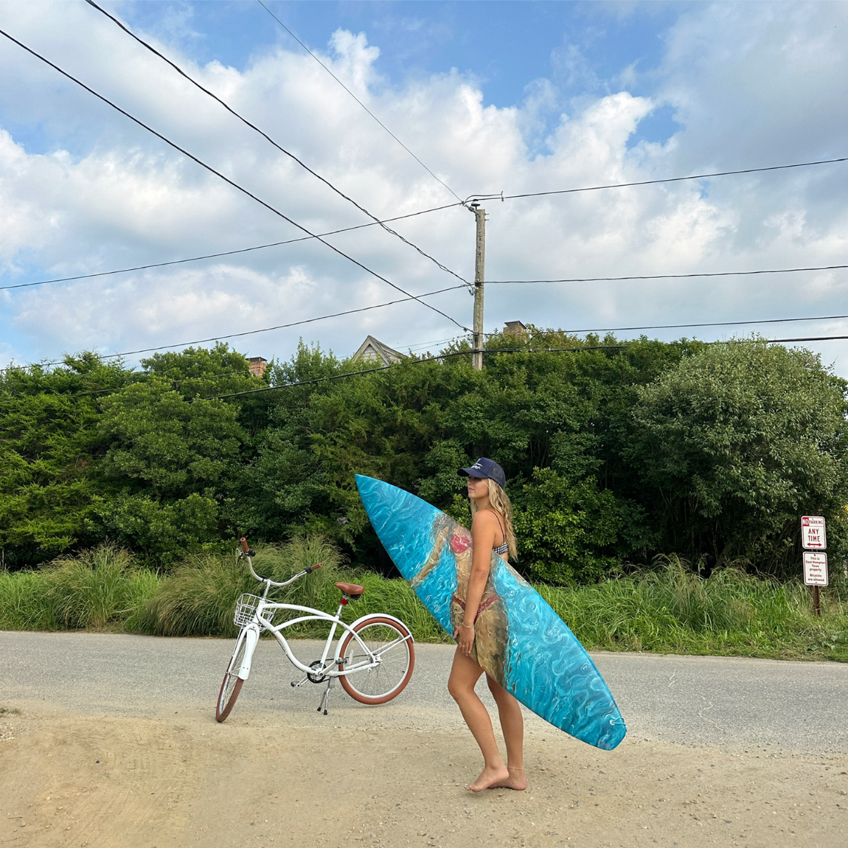 A woman standing on a sandy roadside holding a blue surfboard with a painting of a mermaid. She is barefoot, wearing sunglasses, a baseball cap, and a bikini. A white bicycle with brown tires is on the ground nearby. Behind her there is green foliage, a utility pole, and power lines under a partly cloudy sky.