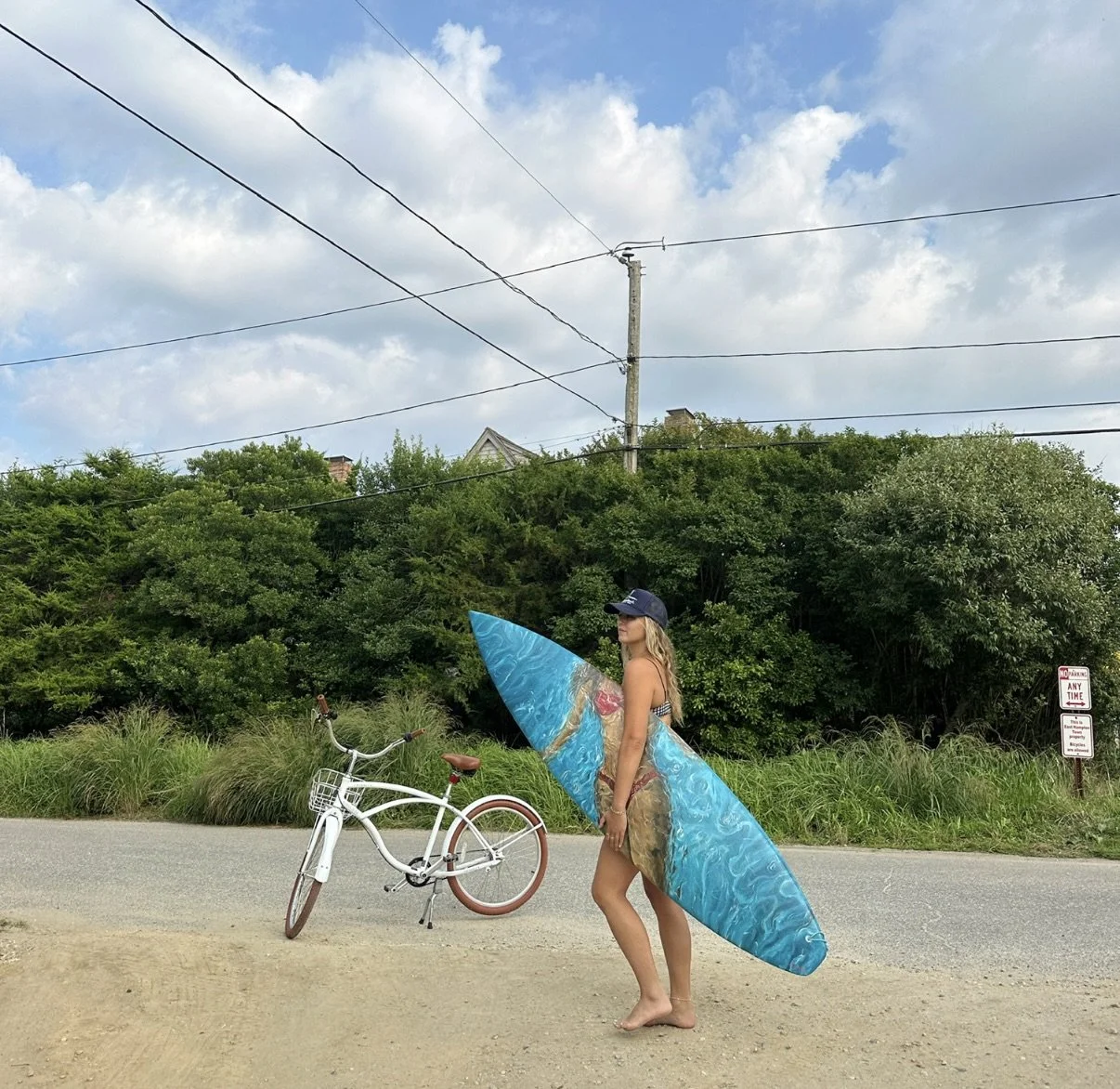 A woman standing barefoot on a gravel area beside a paved road, holding a large blue surfboard. There is a white bicycle with brown tires nearby. In the background, there are green bushes and trees under a partly cloudy sky, with a utility pole and power lines visible.