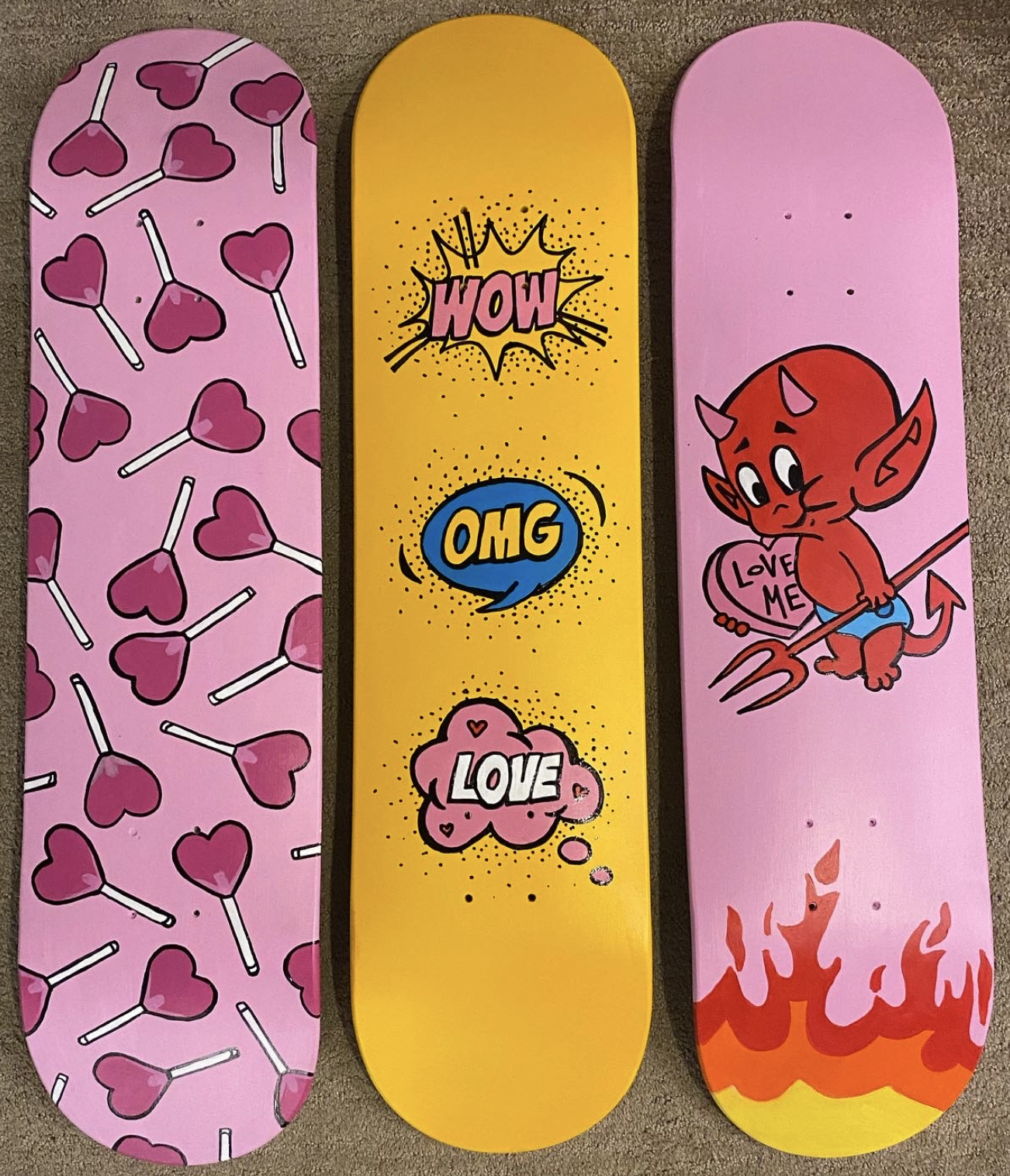 Three skateboard decks with colorful cartoon designs. The left deck has pink background with pink hearts and white lollipop sticks. The middle deck has a yellow background with comic book style words 'WOW', 'OMG', and 'LOVE' in speech bubbles. The ri