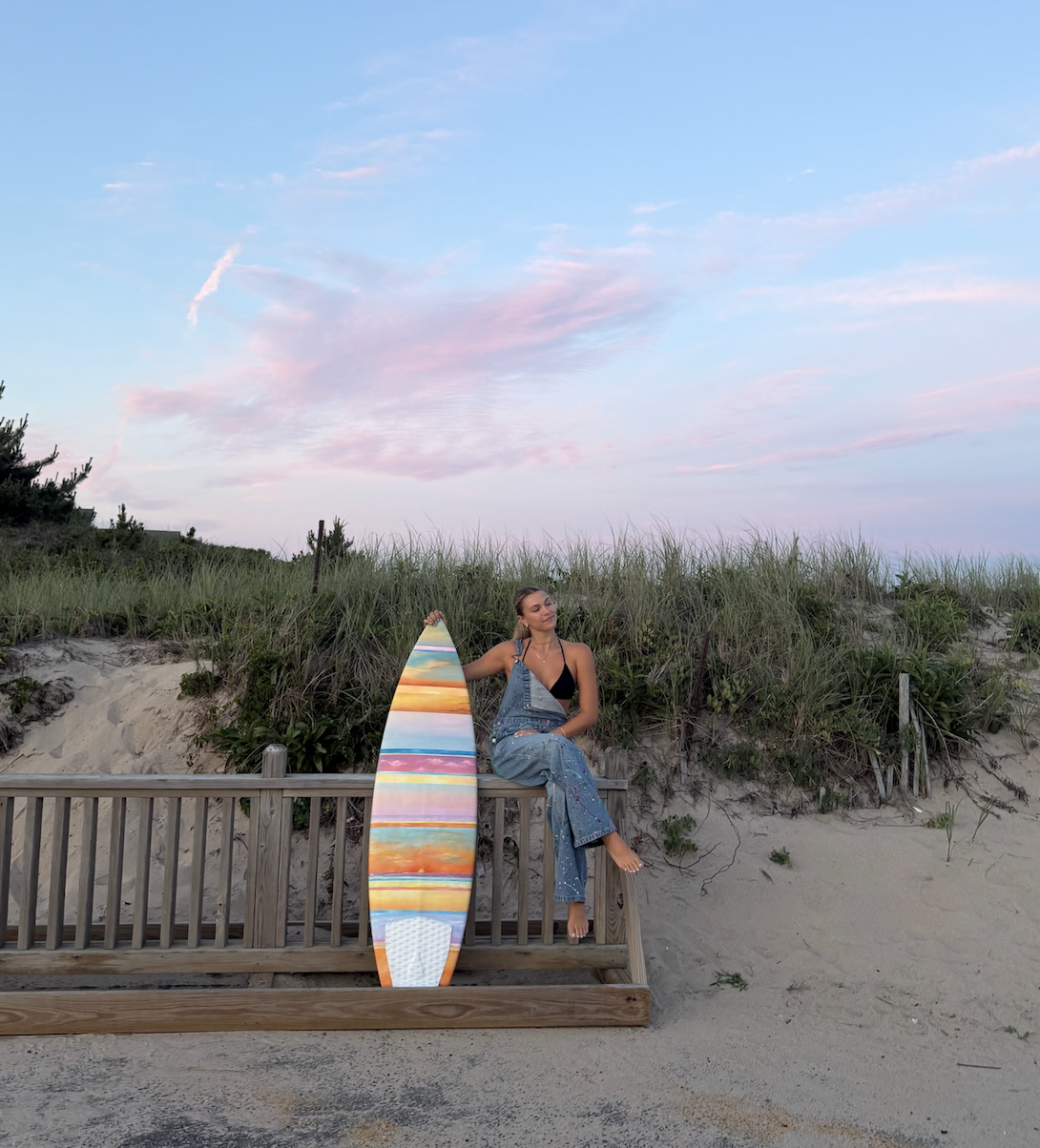 A woman sitting on a wooden railing at the beach holding a colorful striped surfboard. She is dressed in denim overalls and a black top, with her hair tied back. The background features sand dunes, green grass, and a partly cloudy sky at sunset.