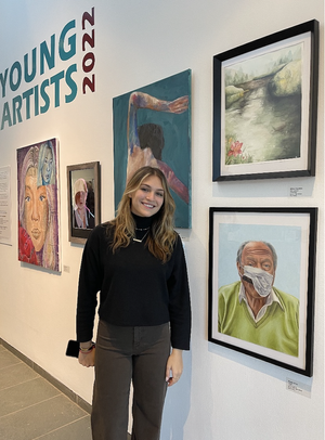 Young smiling woman standing in front of wall with various paintings at an art exhibit.