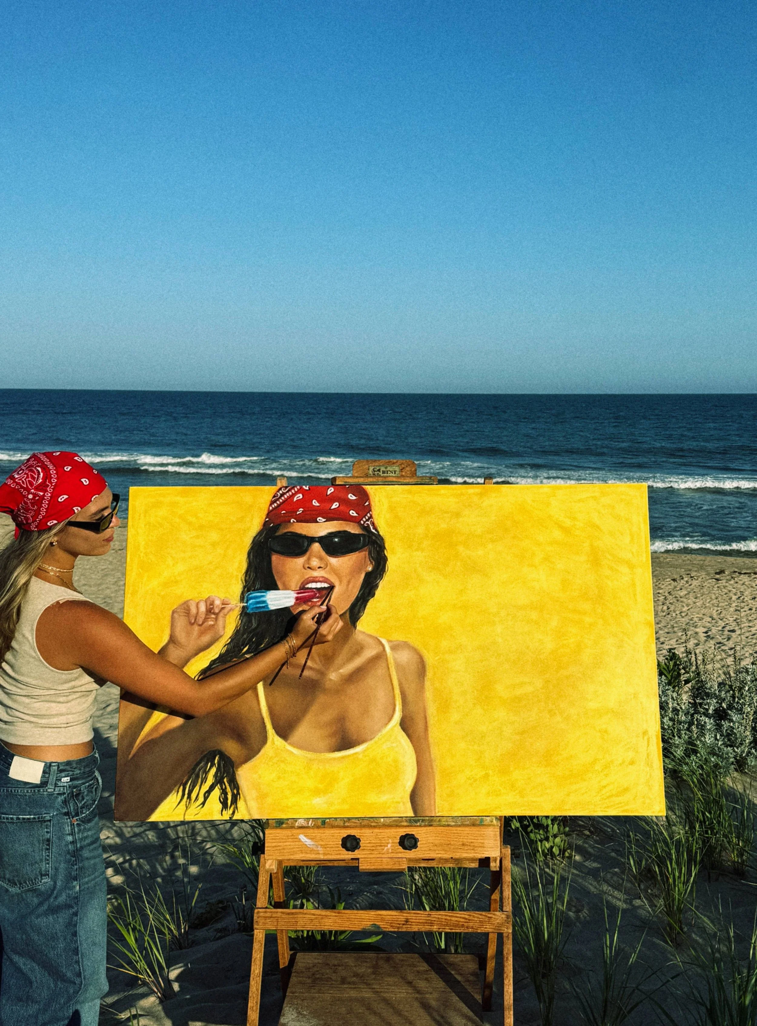 A woman painting a portrait of a woman in yellow swimsuit with sunglasses on a beach. Another woman in a red bandana and sunglasses is assisting with the painting, standing to the left. The scene takes place outdoors near the ocean with a clear sky.