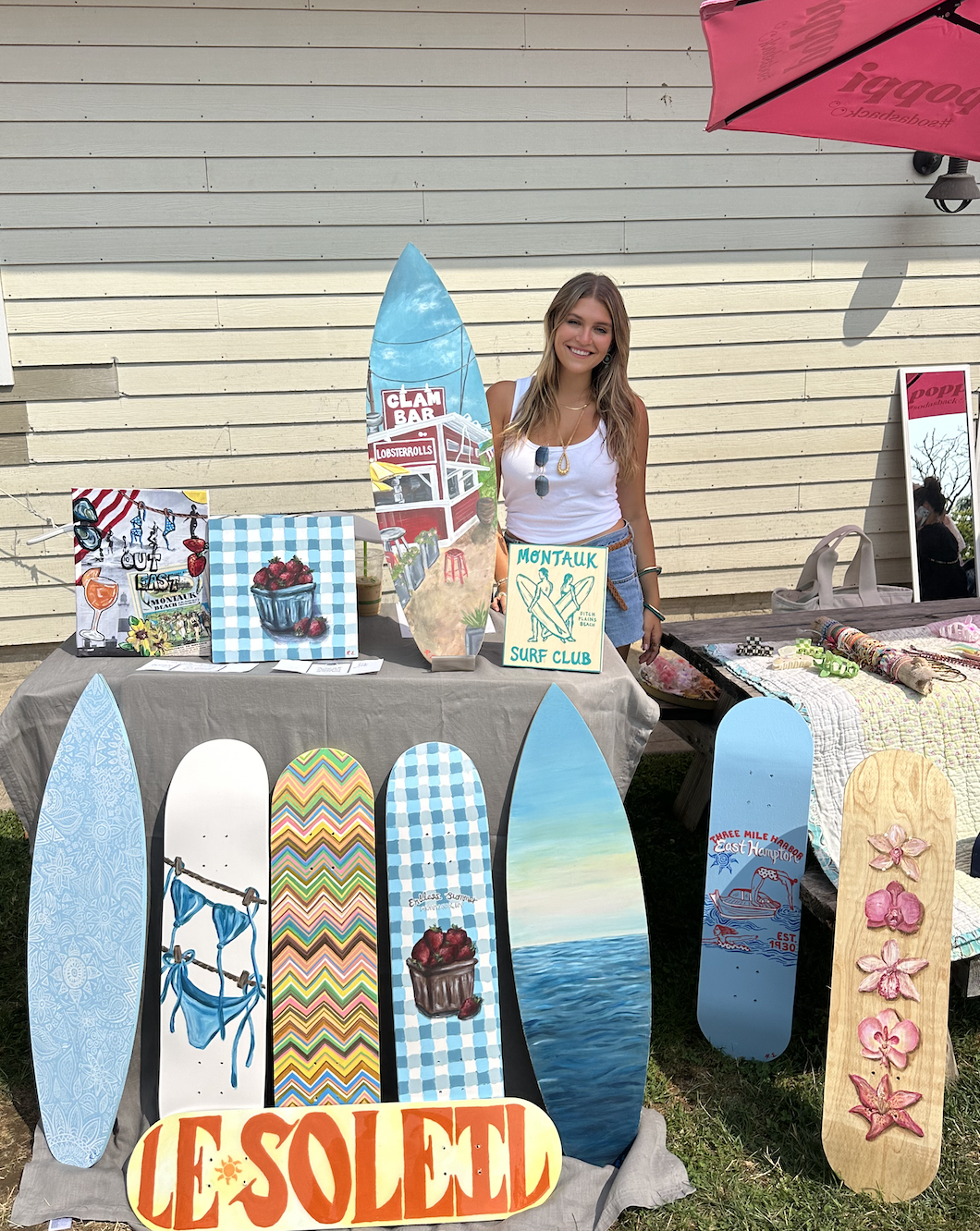 A woman standing behind a table displaying hand-painted surfboards and artwork at an outdoor market. The surfboards feature various designs, including ocean scenes, abstract patterns, and flowers. A sign reads 'Les Sold' and artwork includes a bowl of strawberries and a clam bar scene. The woman is smiling, wearing a white tank top and denim shorts.