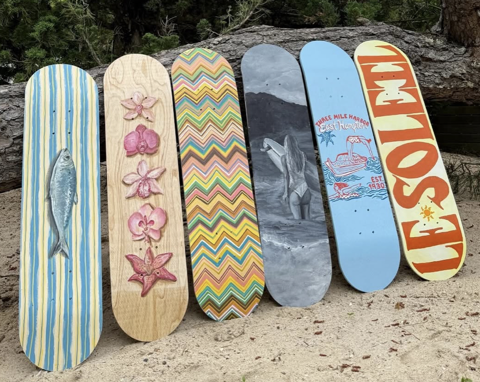 Six skateboards leaning against a tree on a sandy ground, with forest in the background. The skateboards feature a variety of designs, including a fish, pink flowers, colorful zigzags, a black and white surfer girl, a boat scene, and the words 'LIE SO' in bold letters.