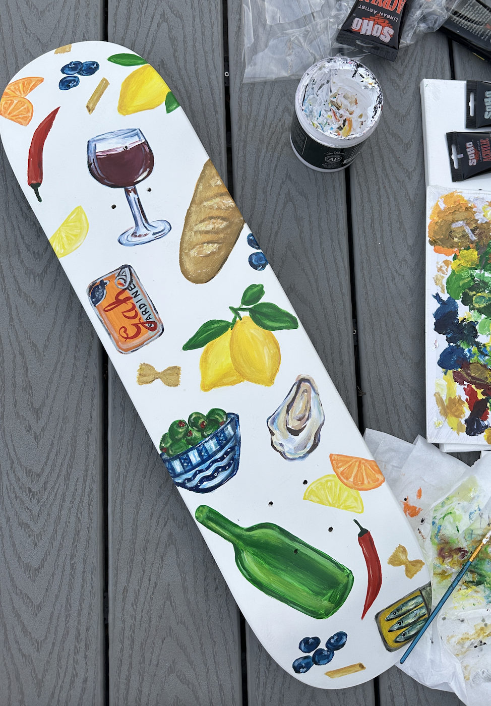 Painted skateboard with colorful food and drink illustrations, including a glass of red wine, lemon, orange, red chili, green bottle, blueberries, loaf of bread, olives, garlic, and lemon.