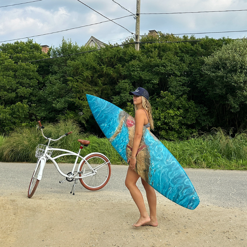 A woman in a bikini, wearing a cap, holding a painted surfboard, stands on the side of a road next to a white bicycle, with green trees and power lines in the background.