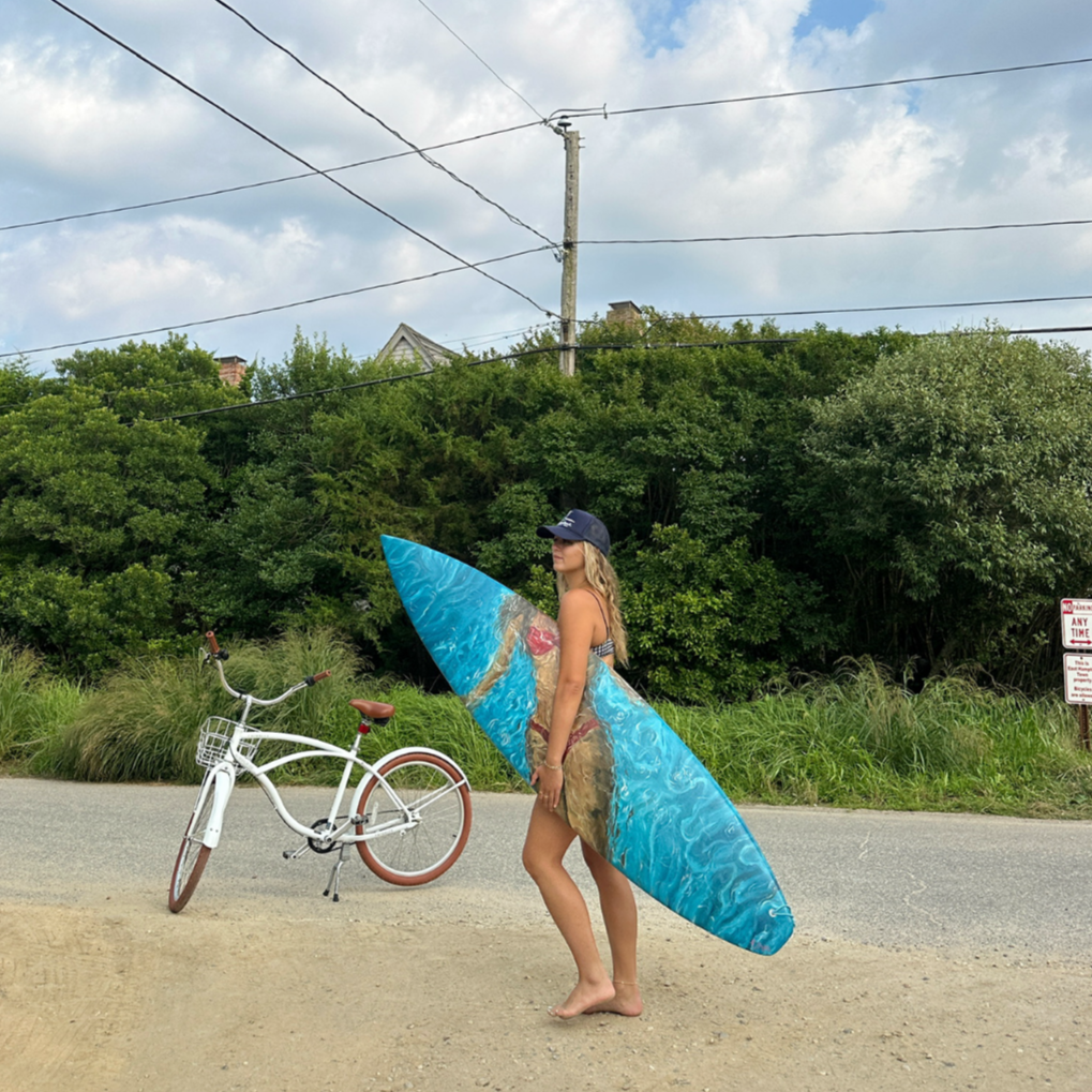 A woman in a swimsuit and a cap holding a surfboard next to a white bicycle on a sandy area with green bushes and trees in the background. Overcast skies are above.