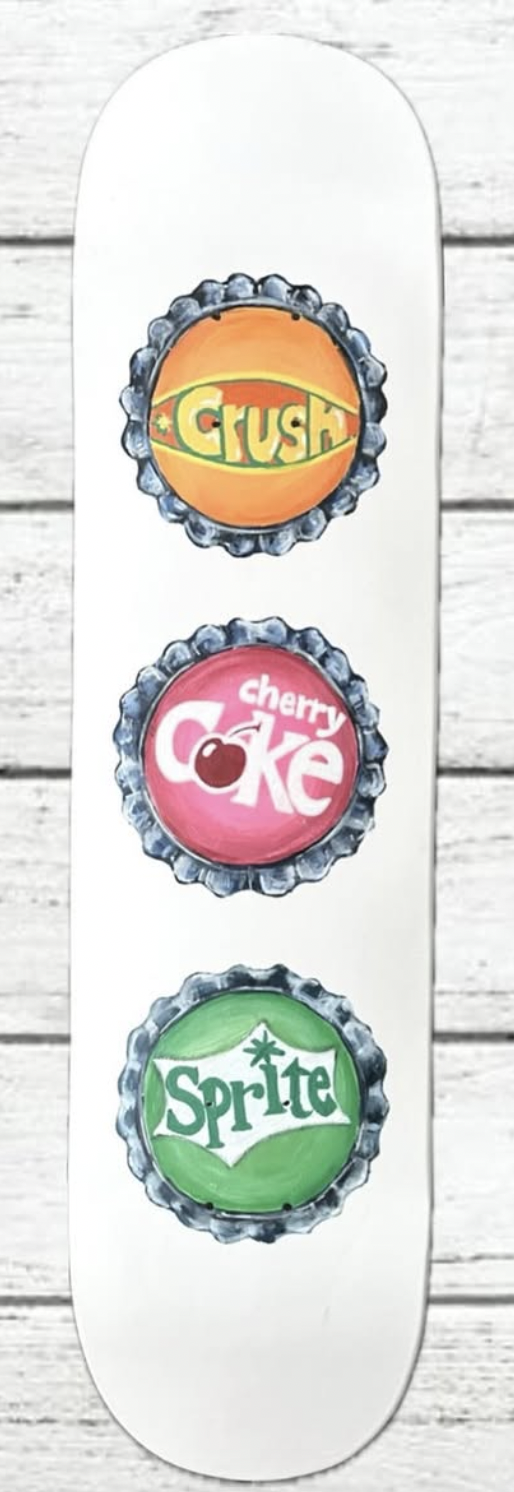 A white skateboard deck with three soda bottle caps painted on it, featuring Crush orange soda, Cherry Coke, and Sprite logos, set against a white wooden background.