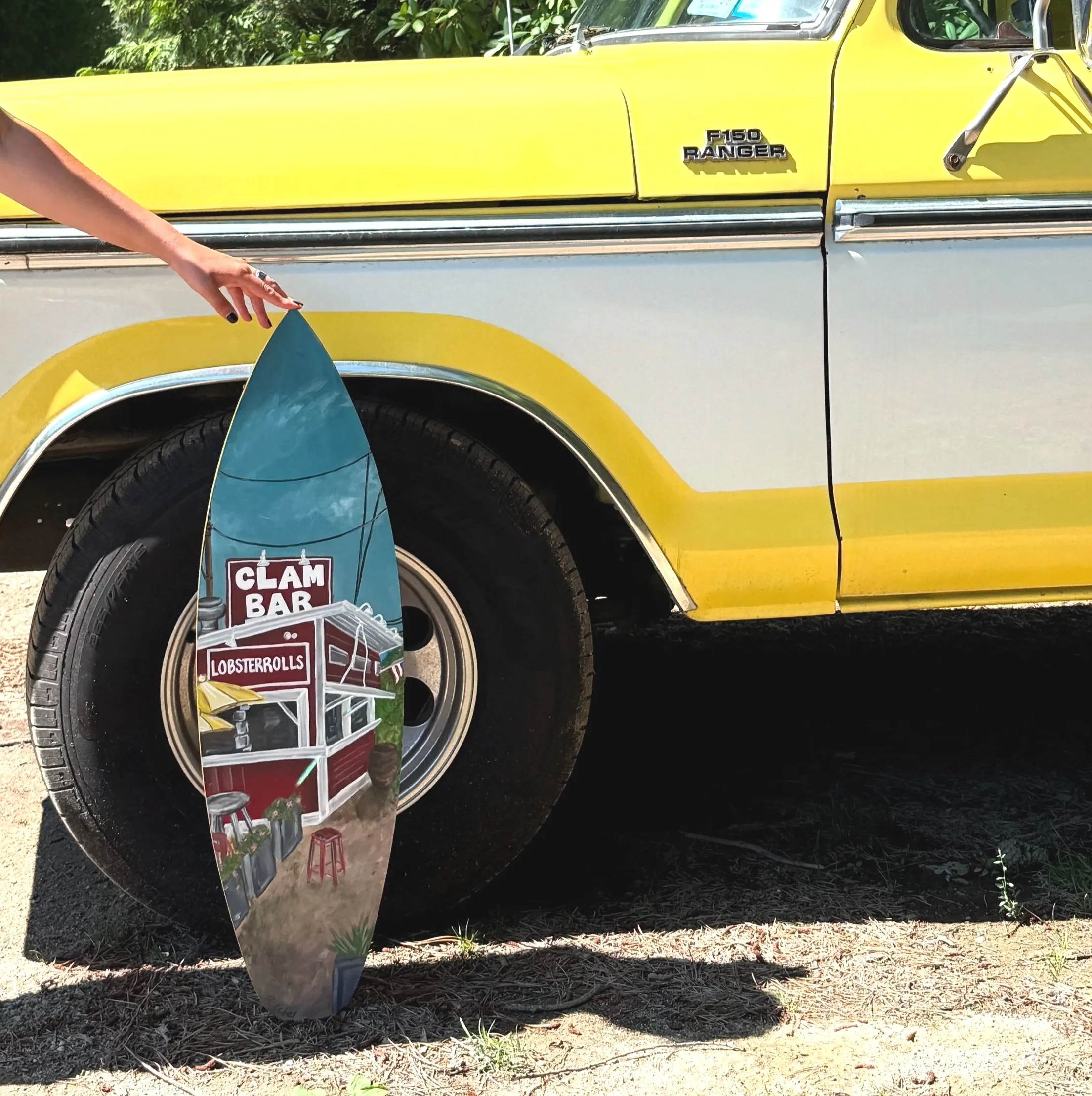 Close-up of a yellow Ford F150 Ranger truck with a part of surfboard leaning against its tire, and a cone-shaped sign advertising a clam bar and lobster rolls placed near the tire.