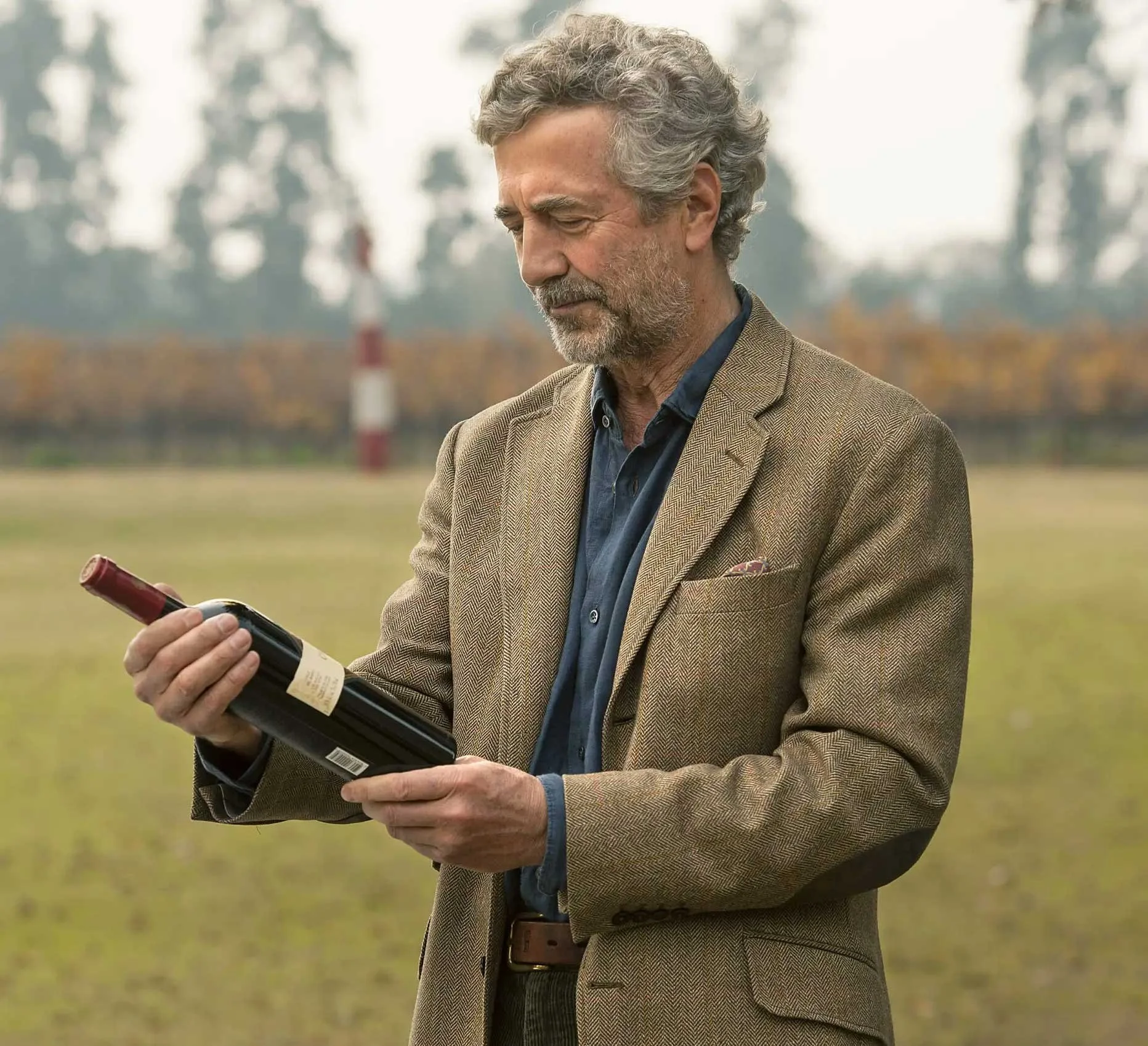 An older man with gray curly hair and a beard, dressed in a brown blazer and navy shirt, holding and looking at a bottle of red wine outdoors on a grassy field with trees in the background.