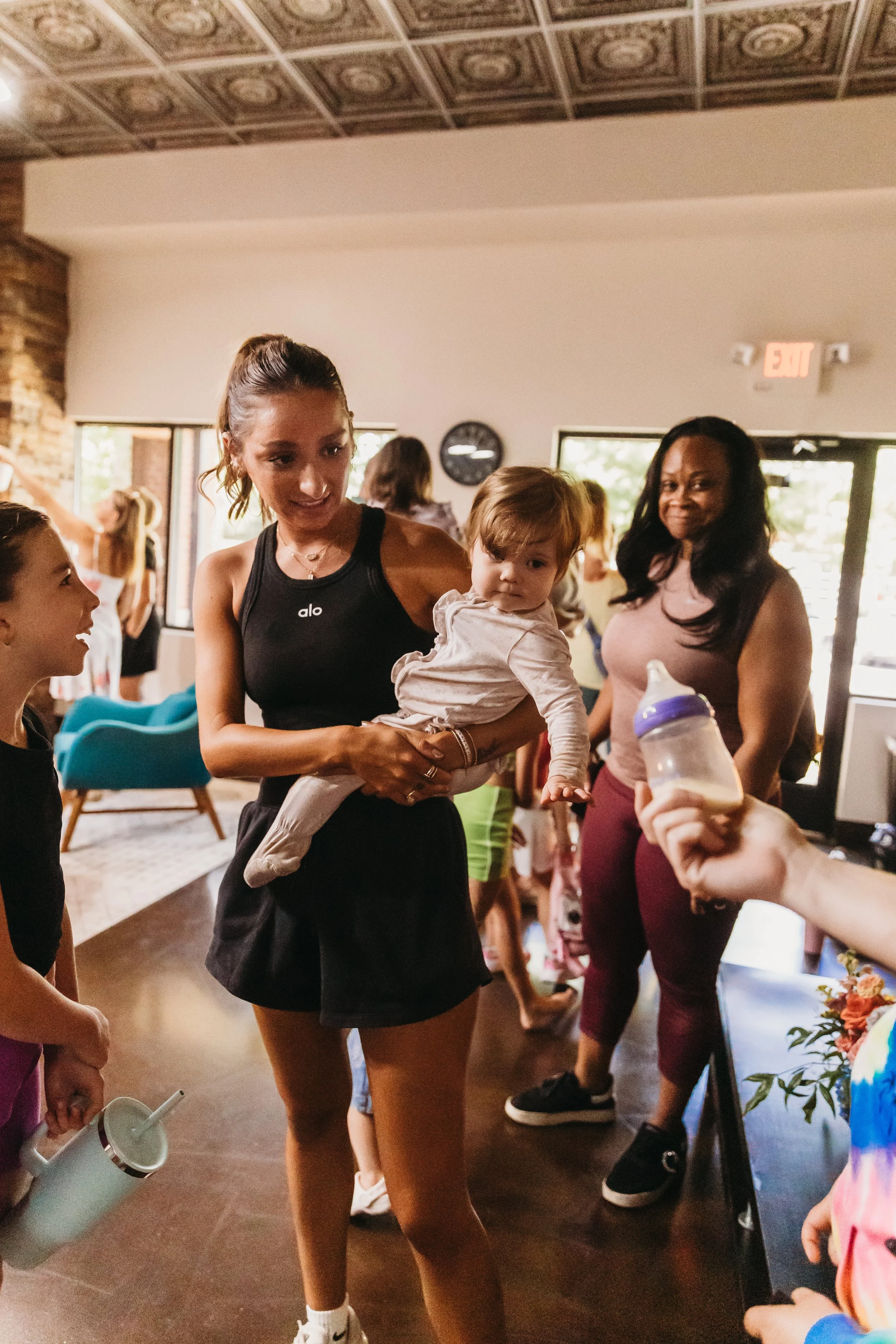 A woman holding a baby and talking to a girl in a room with other people, some holding baby bottles, near a door with an exit sign.