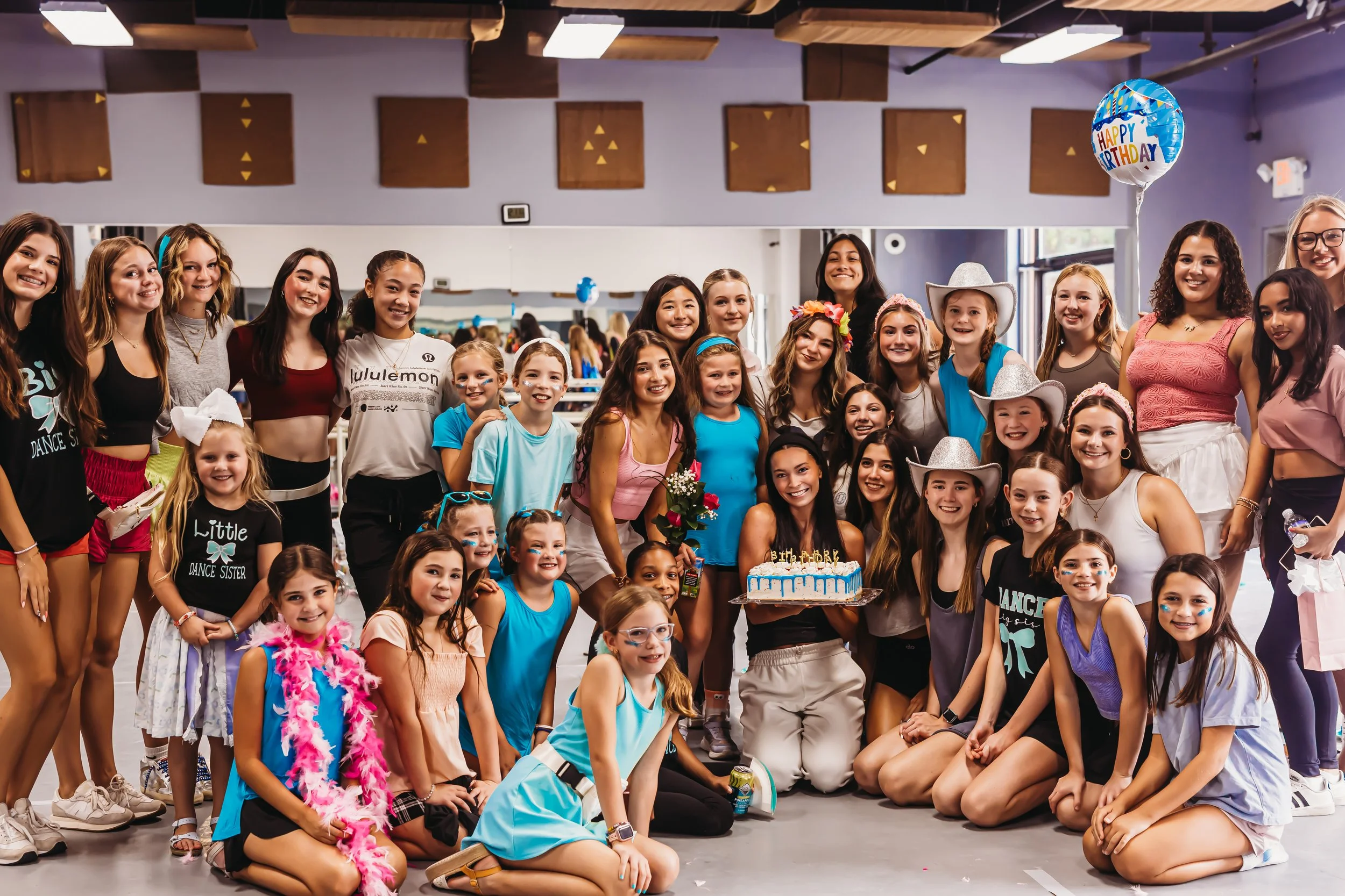 Group of girls and women celebrating a birthday party in a dance studio, with a birthday cake, balloons, and festive decorations.