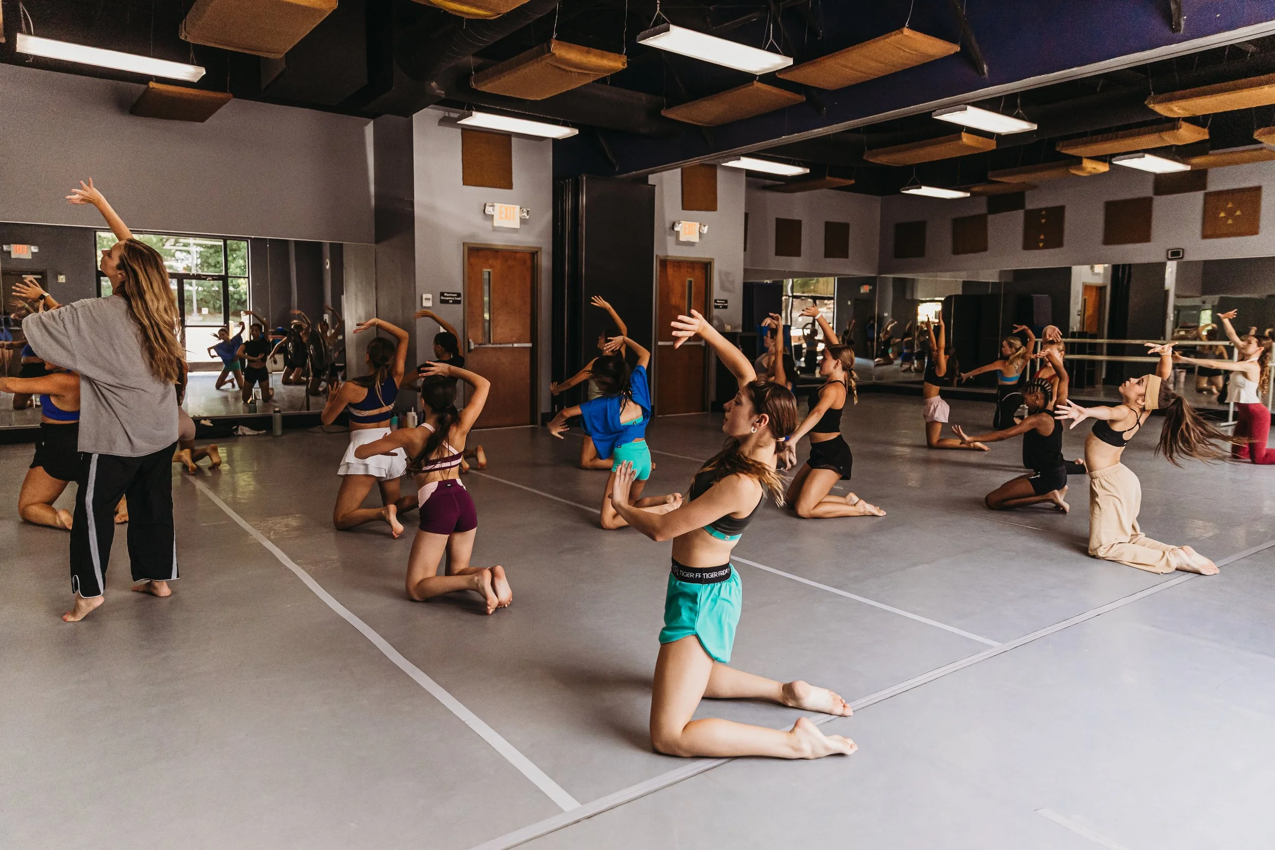 Group of ballet dancers in a dance studio practicing while kneeling on the floor, dressed in workout clothes.