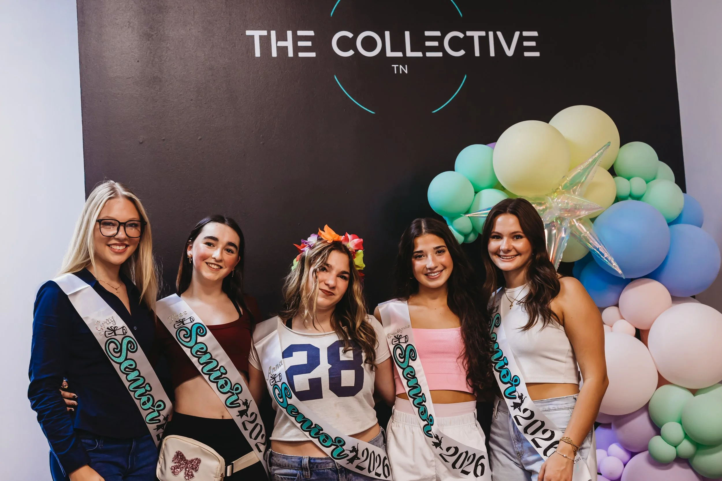 Five young women wearing sashes and graduation gowns at a celebration, standing in front of a backdrop with balloons and a logo that reads 'The Collective'.
