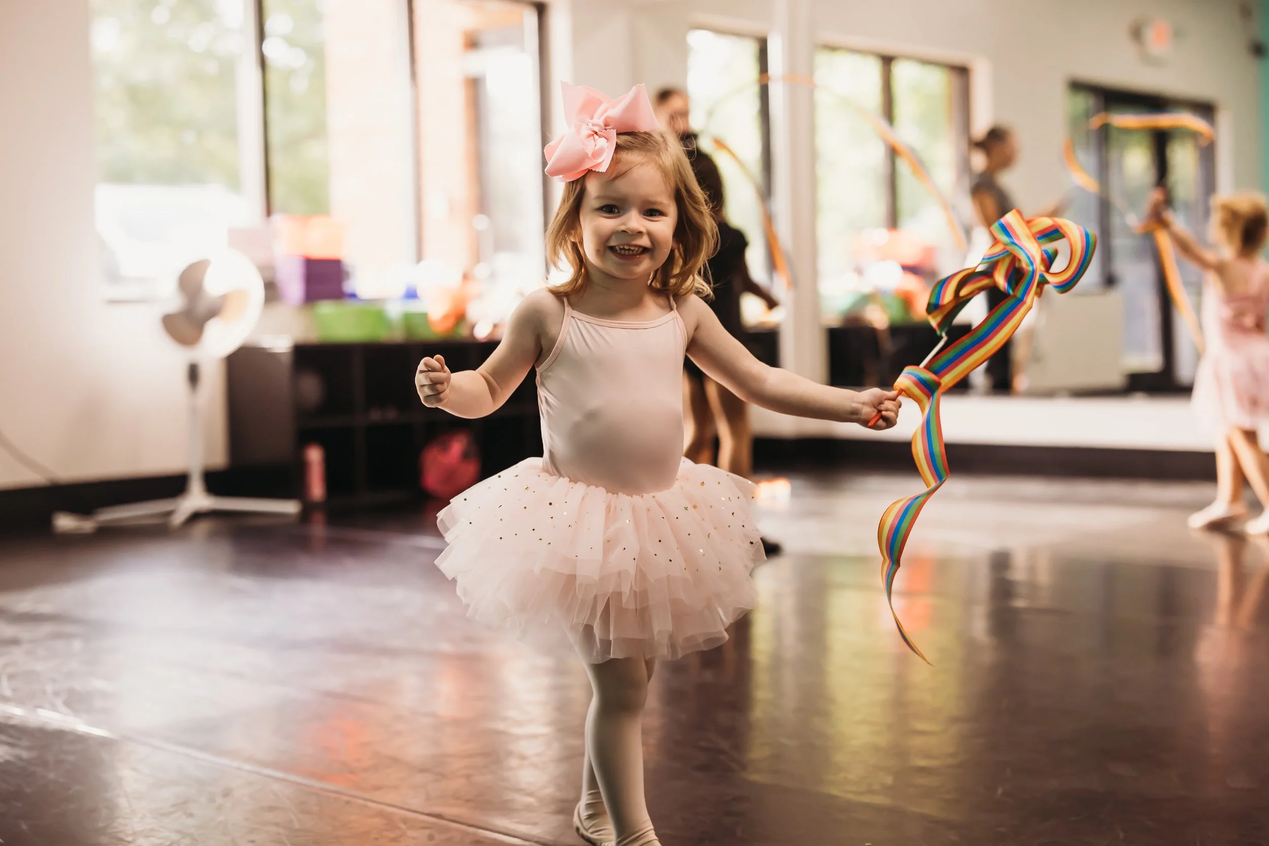 A young girl in a pink tutu dress with a large pink bow in her hair holding a rainbow-colored ribbon, dancing in a party room with other children in the background.