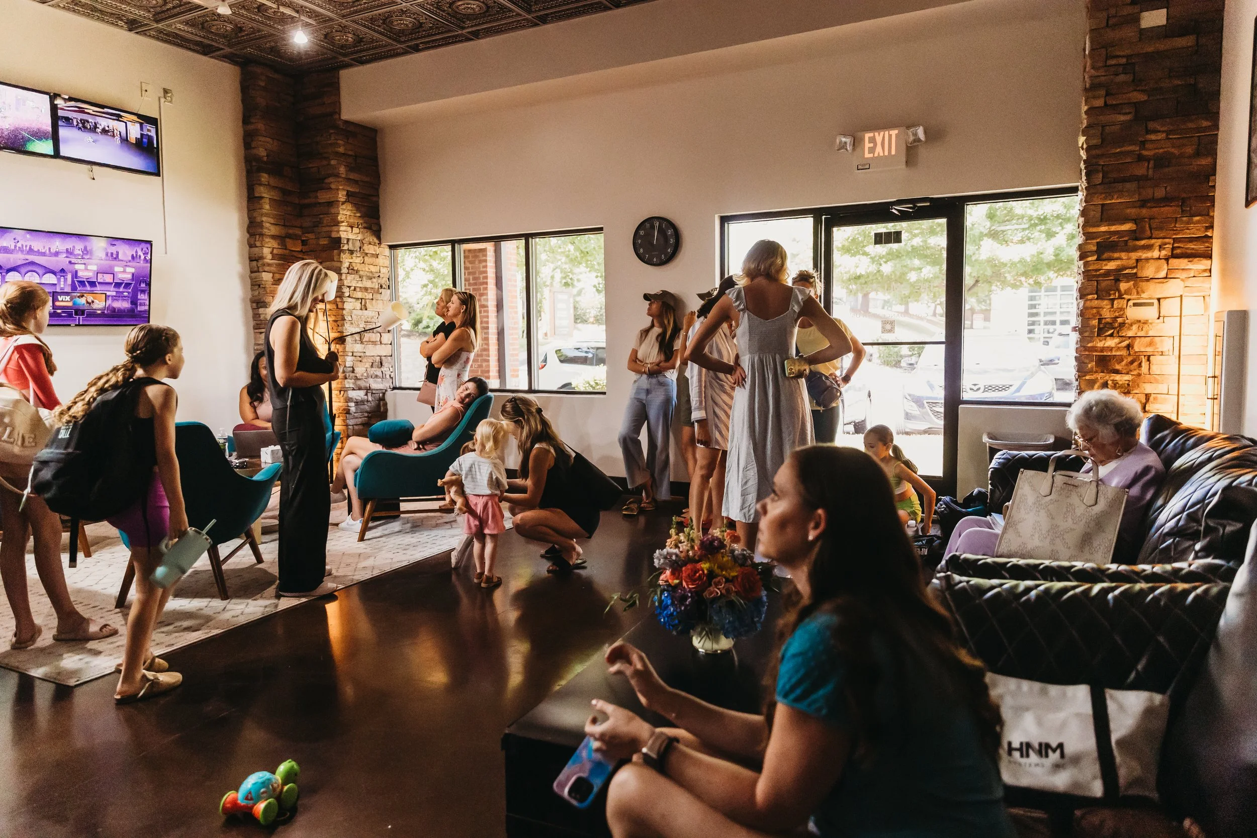 Children and adults in a cozy indoor space, some seated on sofas, others standing or squatting, engaged in various activities, with a large window and TV screens on the wall.