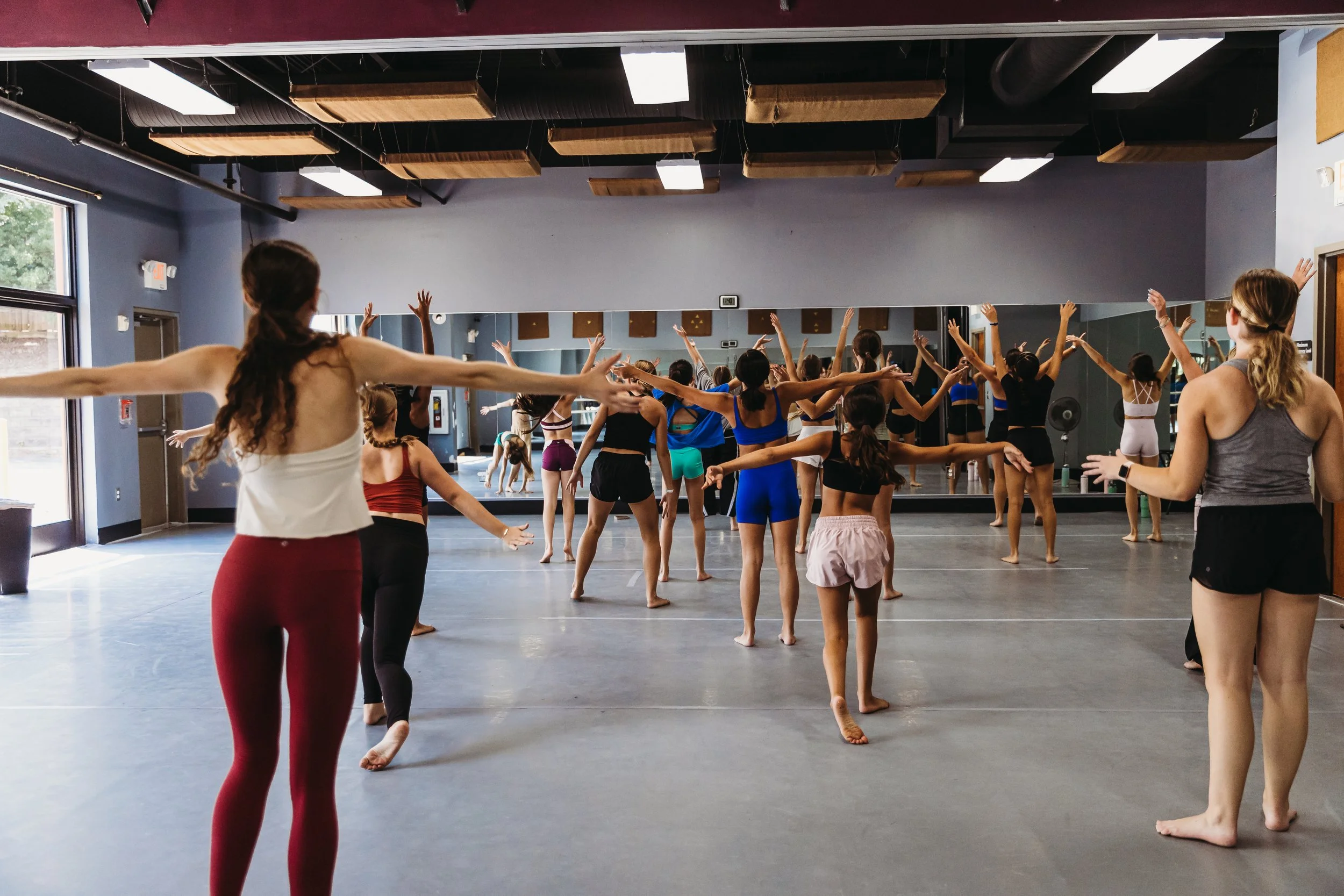 A dance class in a studio with students practicing ballet or contemporary dance, stretching with arms extended, and a mirror reflecting the scene.