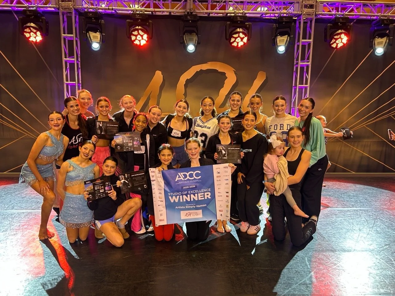 A group of young women and girls celebrating on a stage, holding awards and a large banner that reads 'Studio of Excellence Winner.' They are dressed in dance costumes and casual clothes, with some kneeling in the front and others standing behind. The background features stage lighting and a dark backdrop with gold accents.