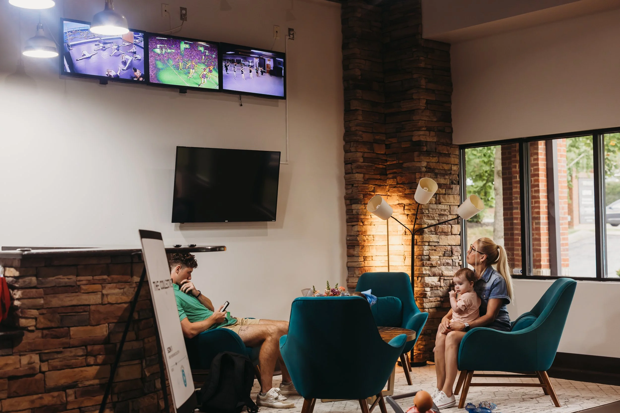 A woman with a young child sitting on her lap in a lounge area, watching TV. A man is seated on a separate chair, looking at his phone. The room has brick accents, large windows, and a modern floor lamp.