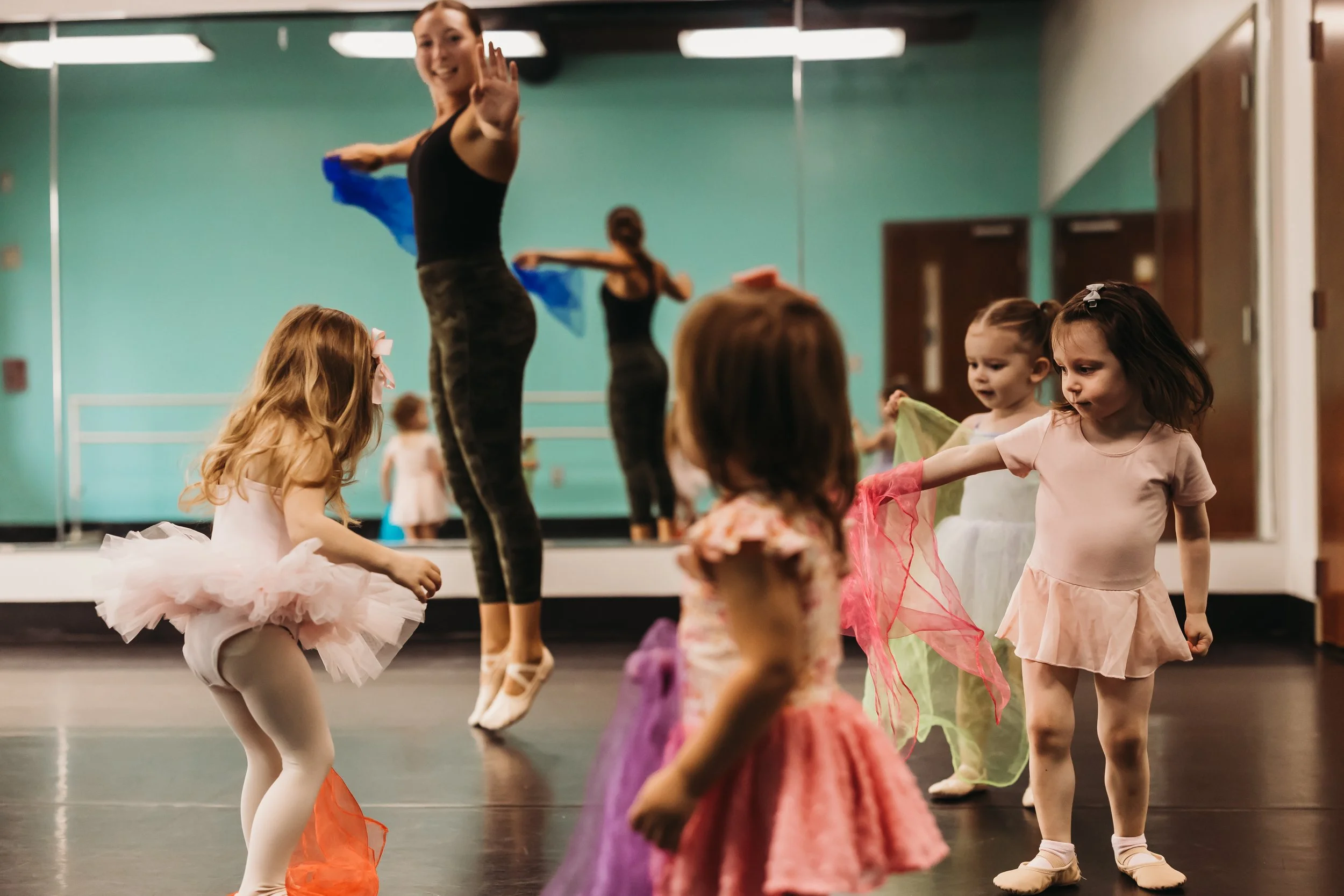 A ballet class with young girls practicing in tutus and a teacher demonstrating in front of a mirror.