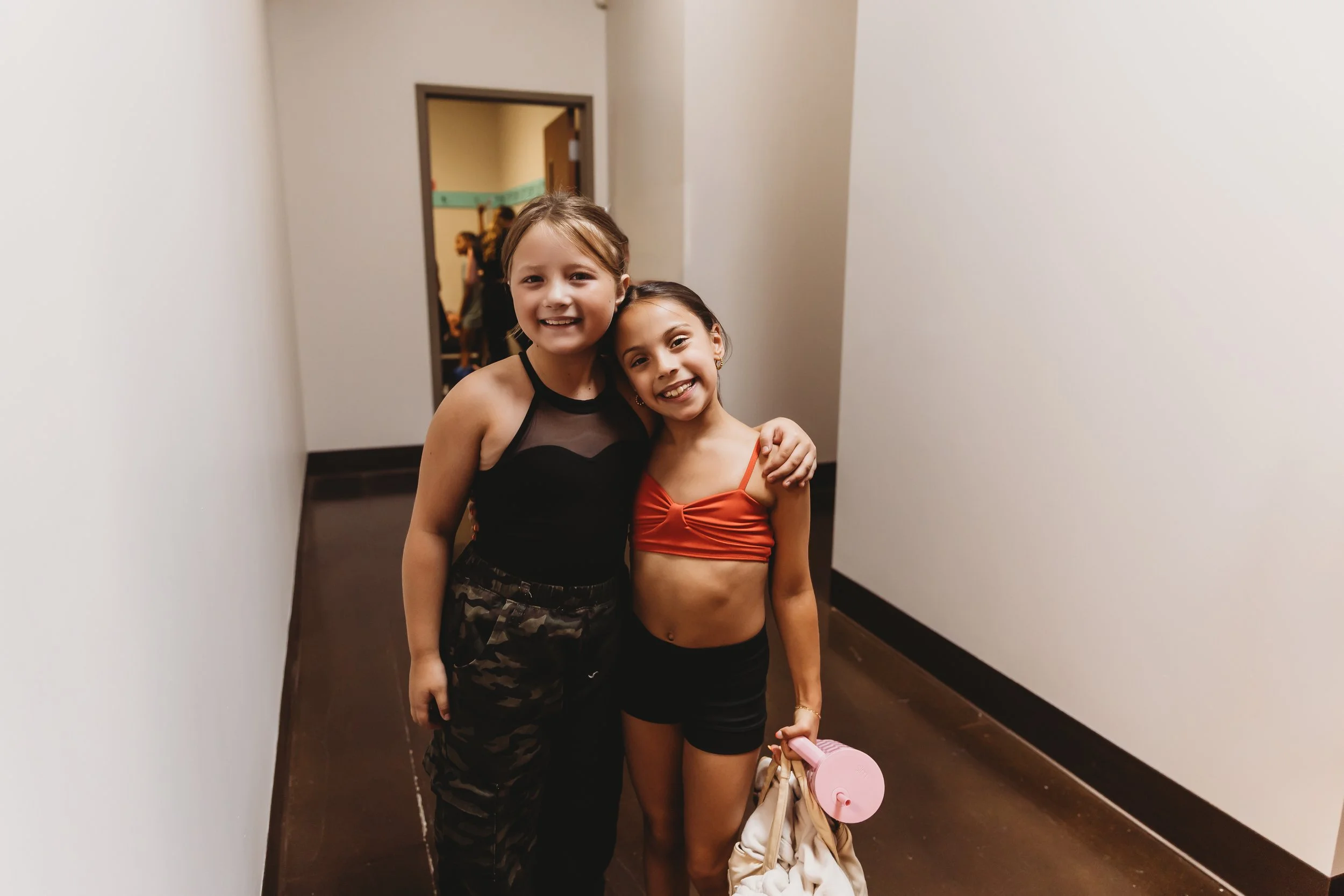 Two young girls standing together in a hallway, smiling at the camera. One girl is wearing a black sleeveless top and camo pants, and the other girl is wearing an orange crop top and black shorts, holding a pink water bottle and a beige bag.