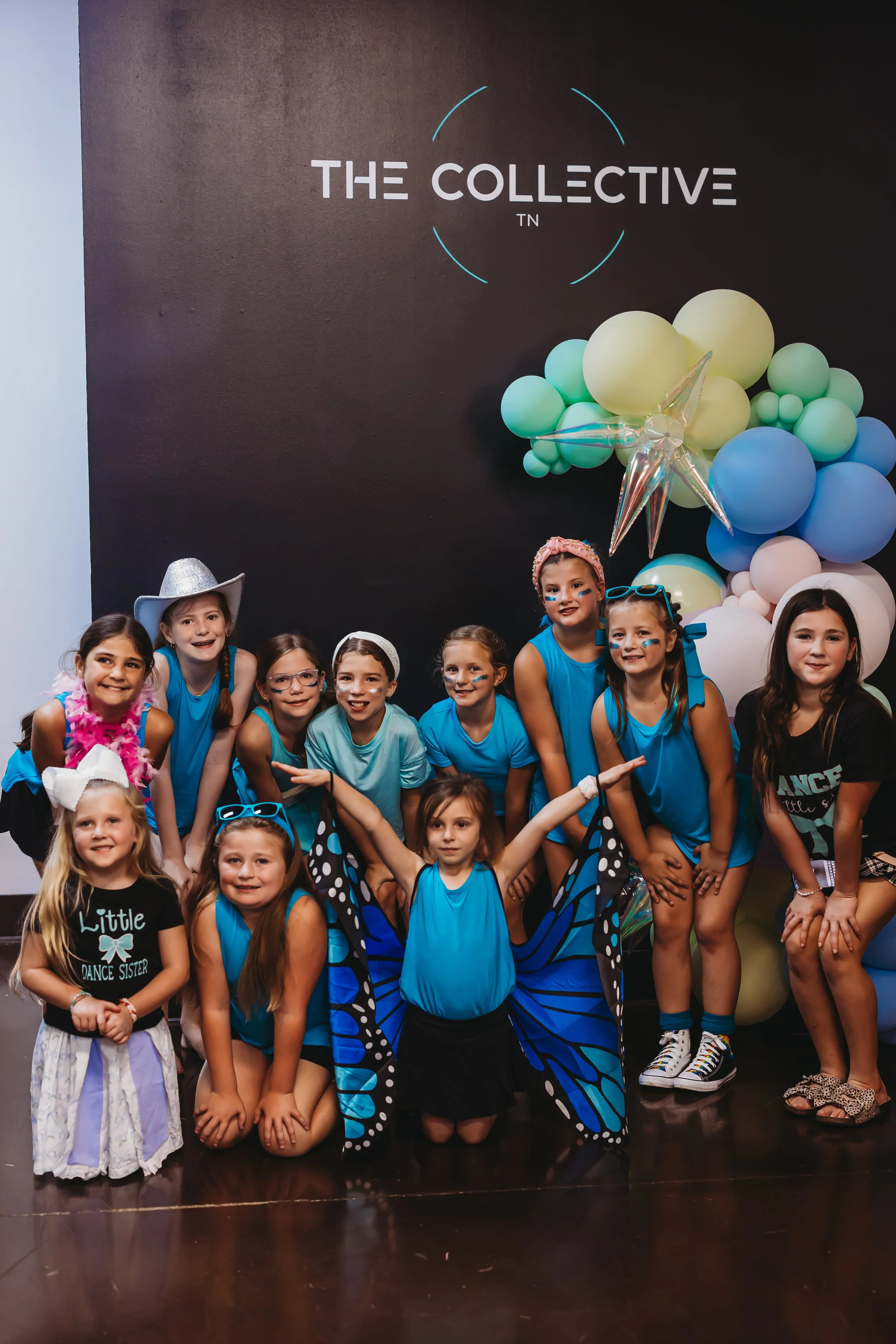 Children dressed in dance costumes pose in front of a black wall with balloons and a star-shaped balloon, with 'The Collective' sign above.