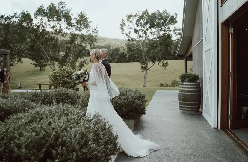 Bride in a white wedding dress holding a bouquet walking beside a man in a suit outdoors near a barn, with trees and a grassy landscape in the background.