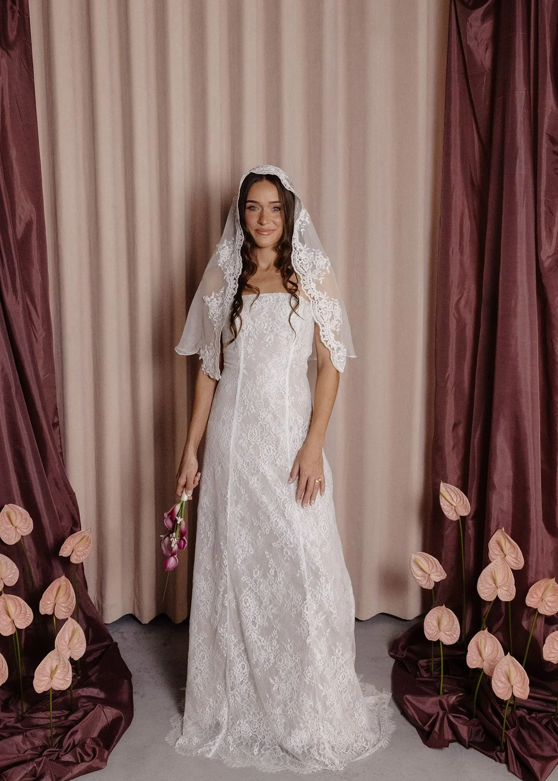 A woman in a white dress holding a bouquet of white flowers, standing against a plain background with greenery above.