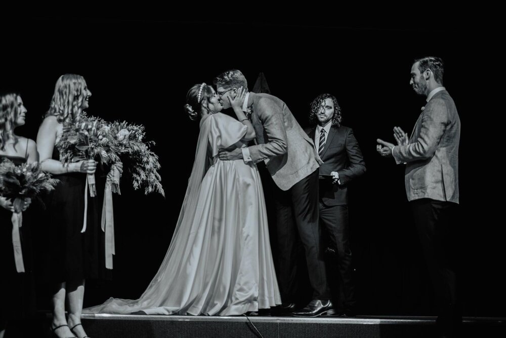 Black and white photo of a wedding ceremony where a bride and groom share a kiss while standing on stage, surrounded by bridesmaids holding bouquets and a man in a suit officiating.
