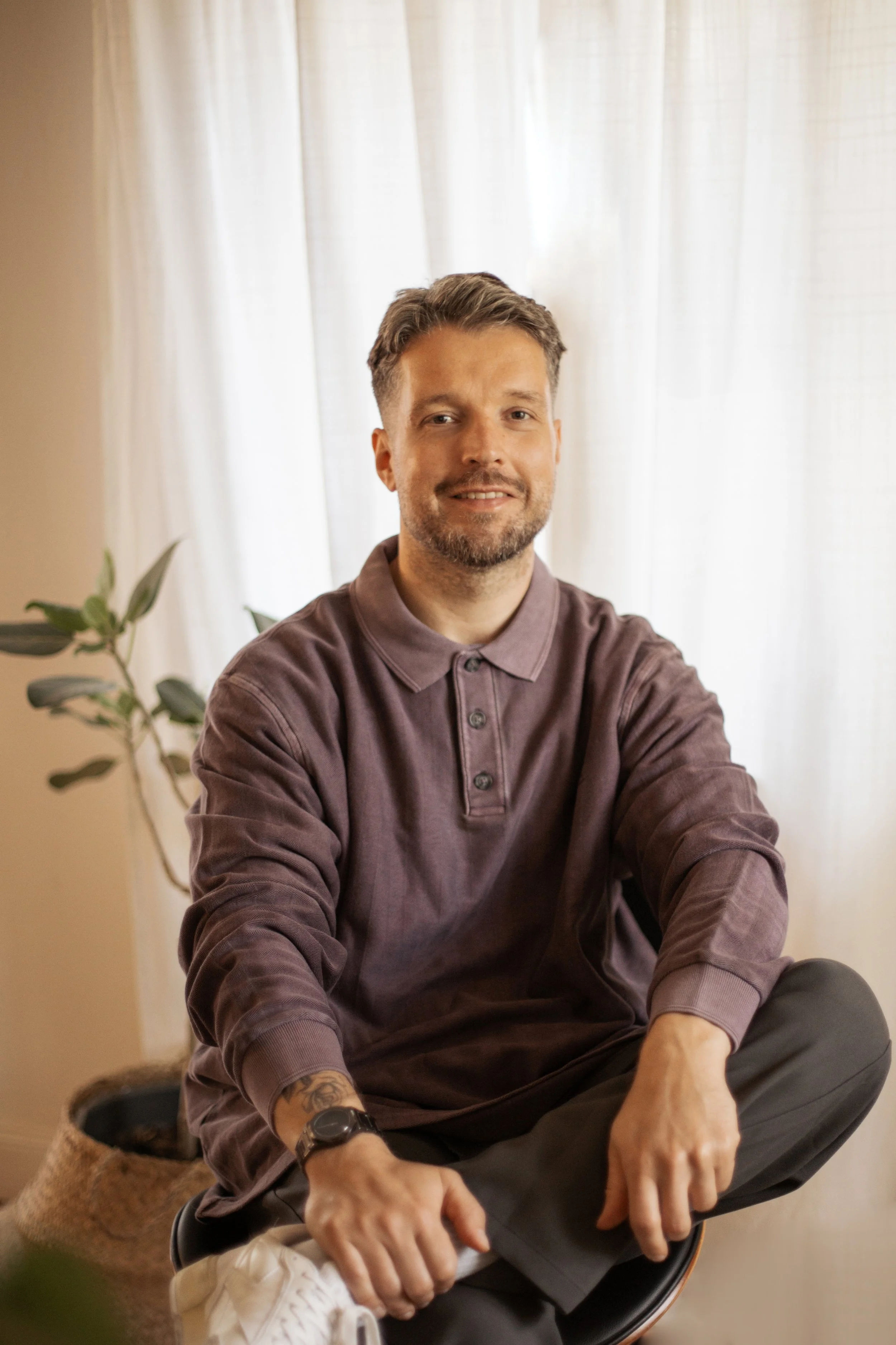 Ein Mann sitzt entspannt in einem gemütlichen Raum vor einem weißen Vorhang und lächelt. Er trägt ein braunes Langarmshirt und eine Armbanduhr.