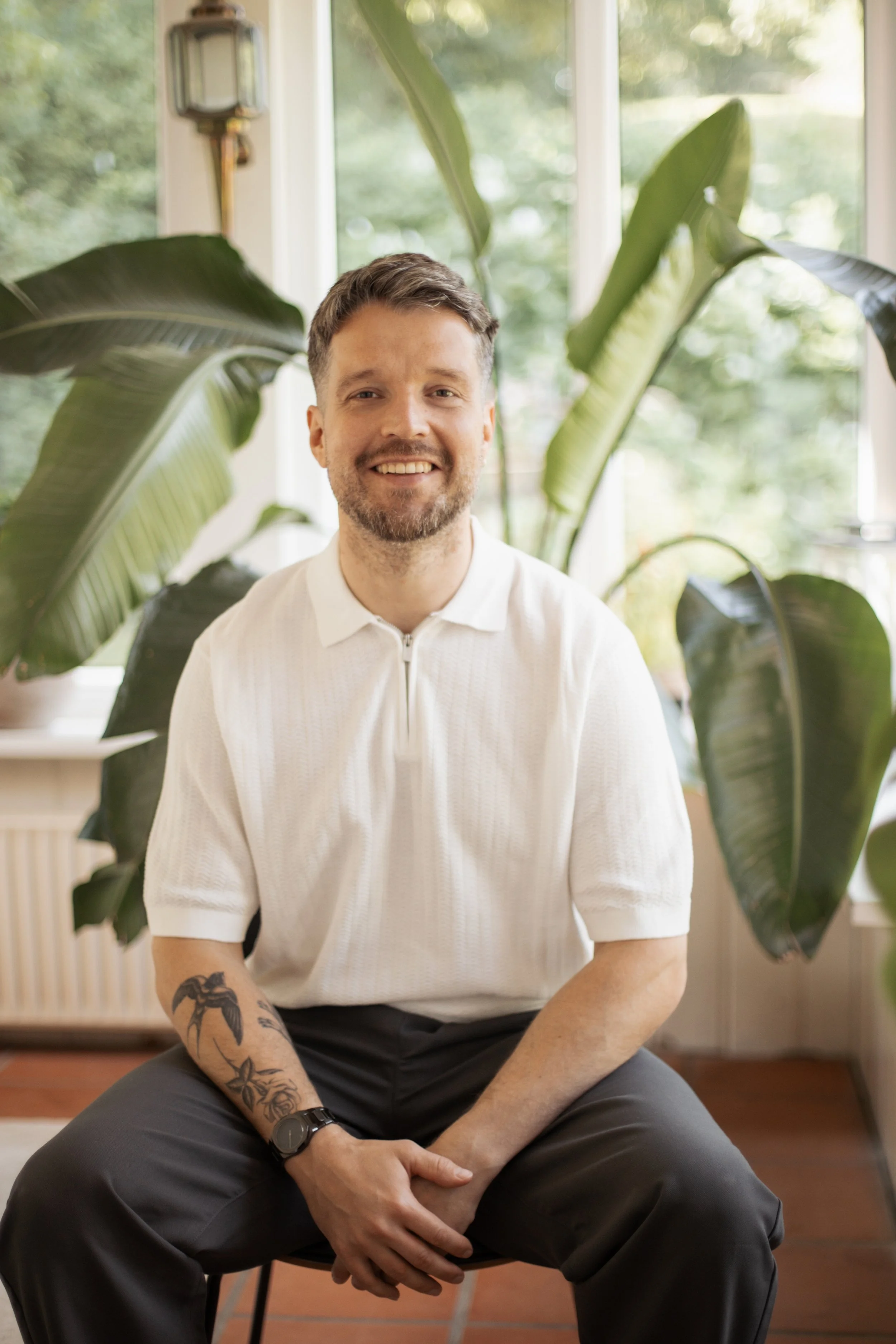 Ein lachender Mann mit Bart und Tattoos am rechten Unterarm sitzt vor großen Zimmerpflanzen und einem großen Fenster, das natürliches Licht hereinlässt, in einem hellen Raum.