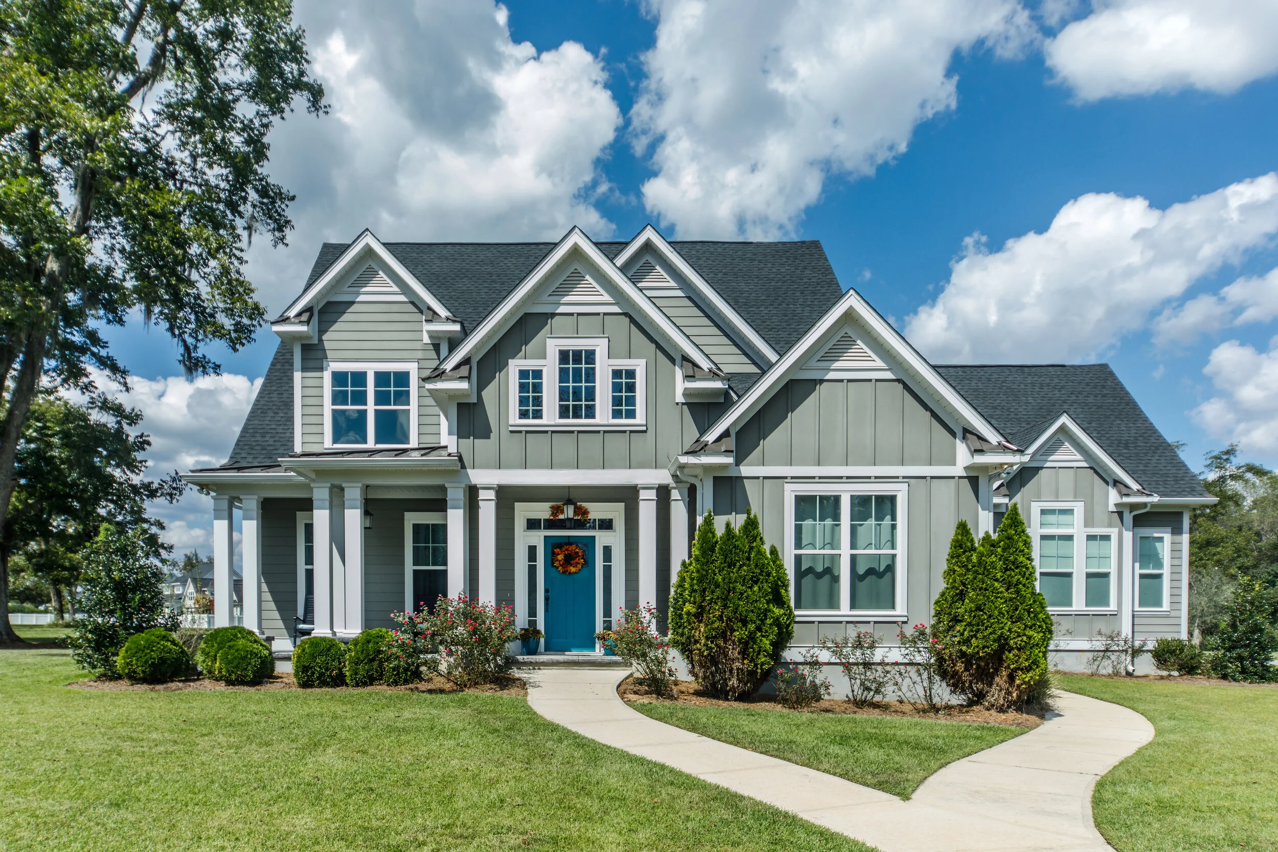 A large, gray, two-story house with white trim and a black shingle roof, decorated for fall with a wreath on the front door, surrounded by a well-maintained lawn and bushes, under a partly cloudy sky.