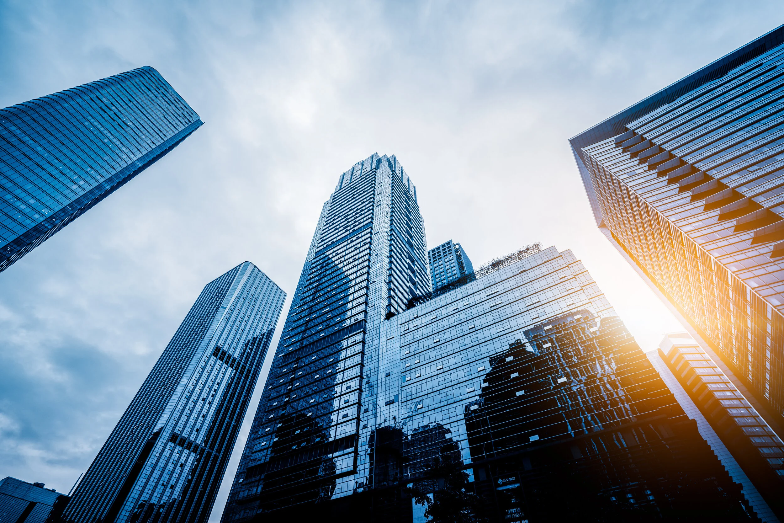 Low-angle view of modern glass skyscrapers reflecting the sky with sun shining behind them, during daytime.