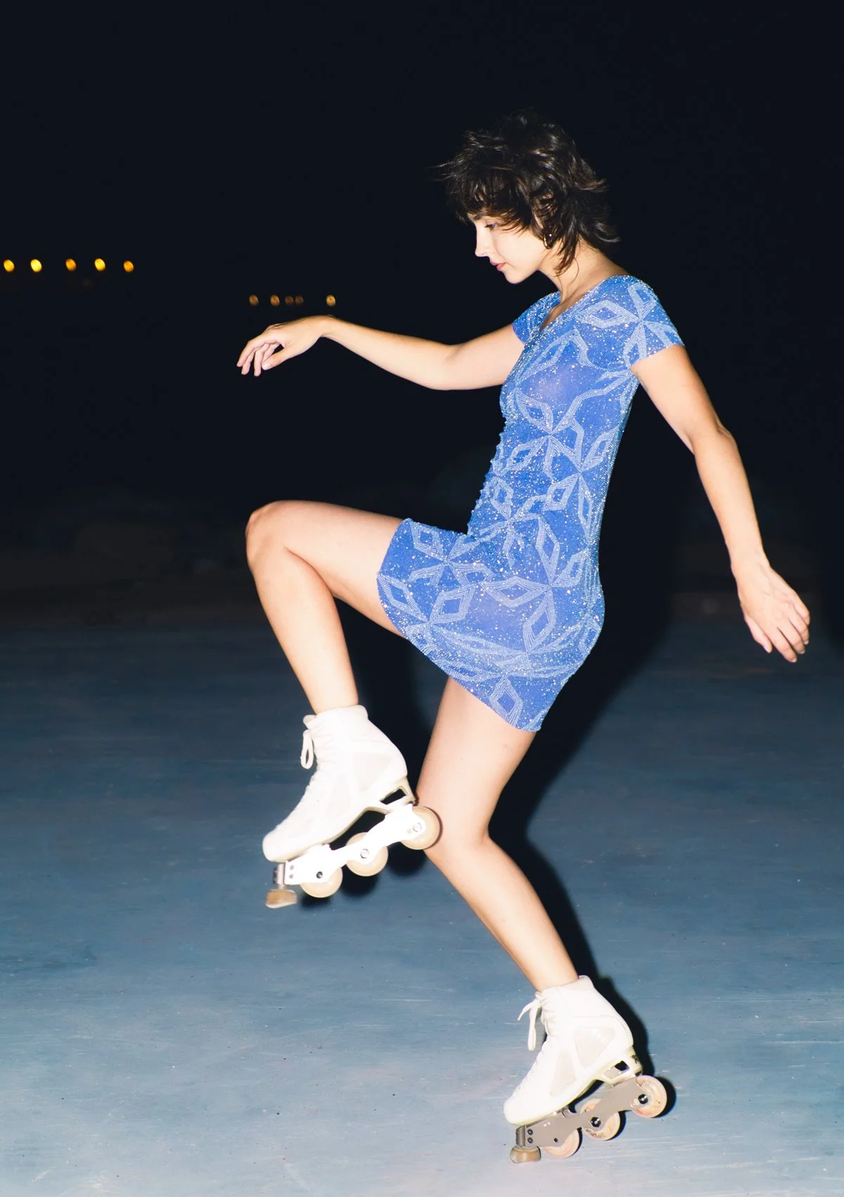 Joven patinando en patines sobre hielo en la noche, vestida con un vestido azul y cabello rizado.