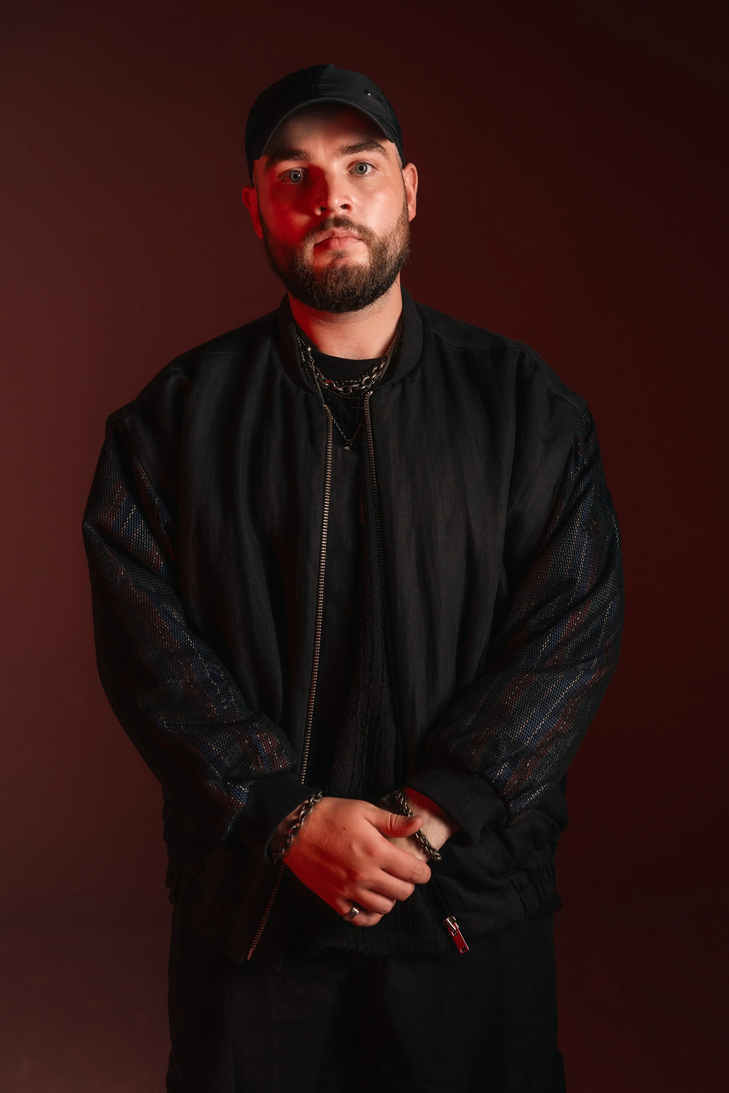 Hombre con barba y gorra negra, vestido con chaqueta negra, posando con fondo oscuro.