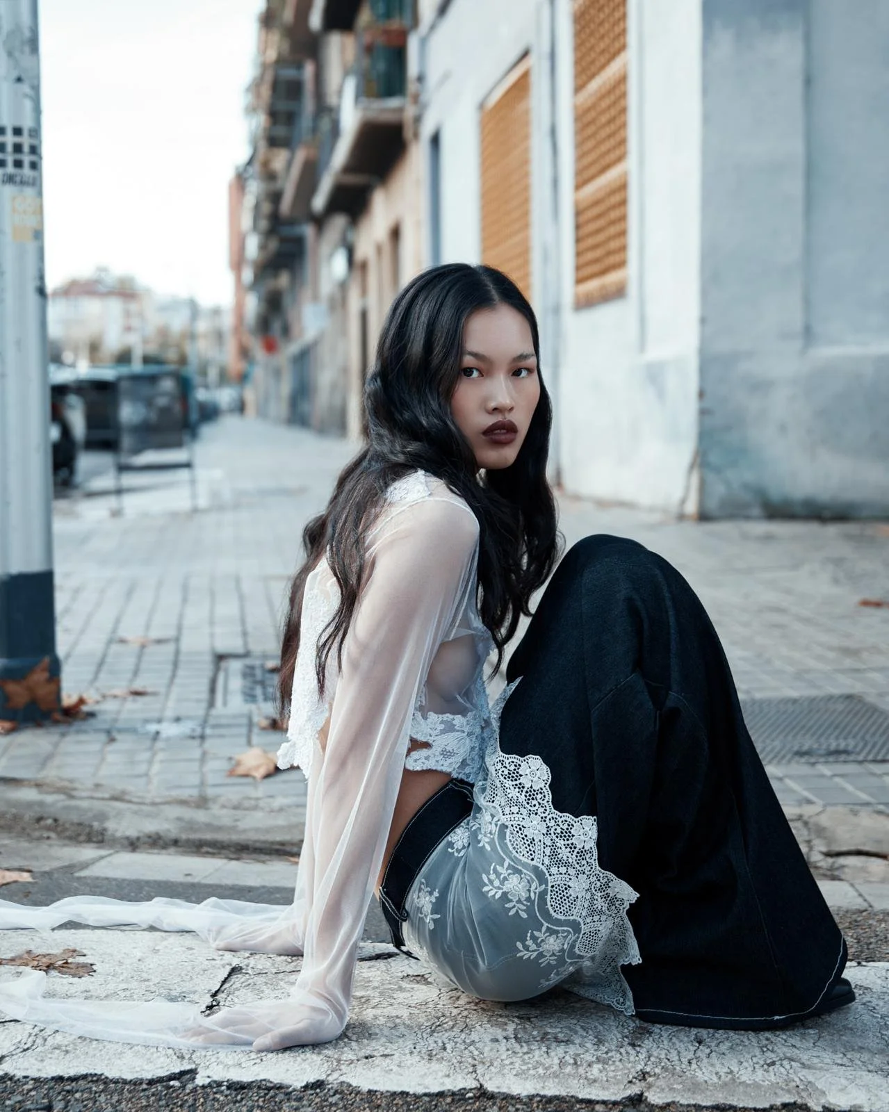 Mujer de cabello oscuro y labios oscuros, con ropa blanca con encaje y pantalones oscuros, sentada en la acera de una calle urbana, mirando hacia la cámara.
