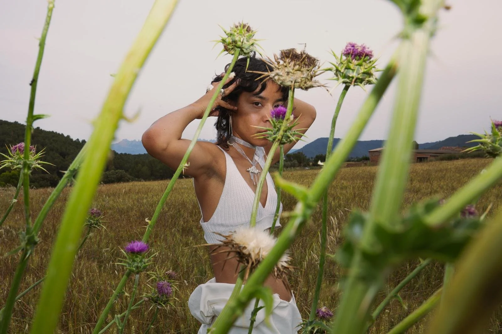 Joven en un campo de flores moradas, viste una blusa blanca y accesorios de joyería, con las manos en la cabeza, rodeada de plantas altas y un paisaje montañoso en el fondo.
