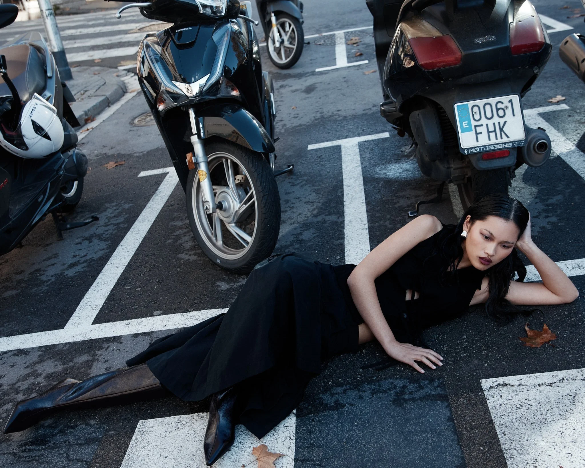 Una mujer vestida de negro acostada en el suelo de un estacionamiento, cerca de motocicletas y un scooter, con expresión de sorpresa o angustia.